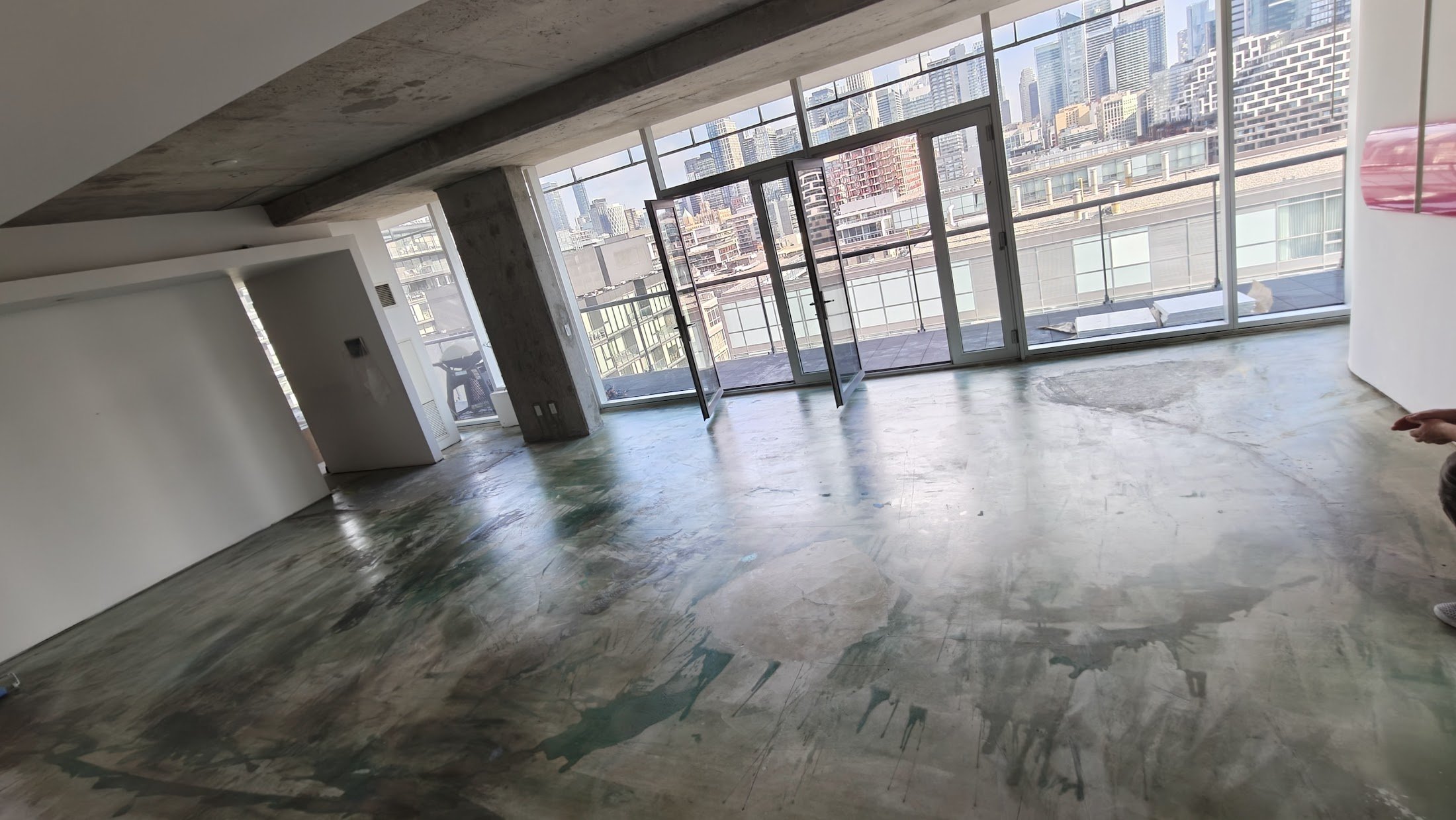 An unfinished high-rise apartment with large floor-to-ceiling windows showing a city skyline. The floor is concrete with patches of paint or material, and there is an open balcony door leading outside.