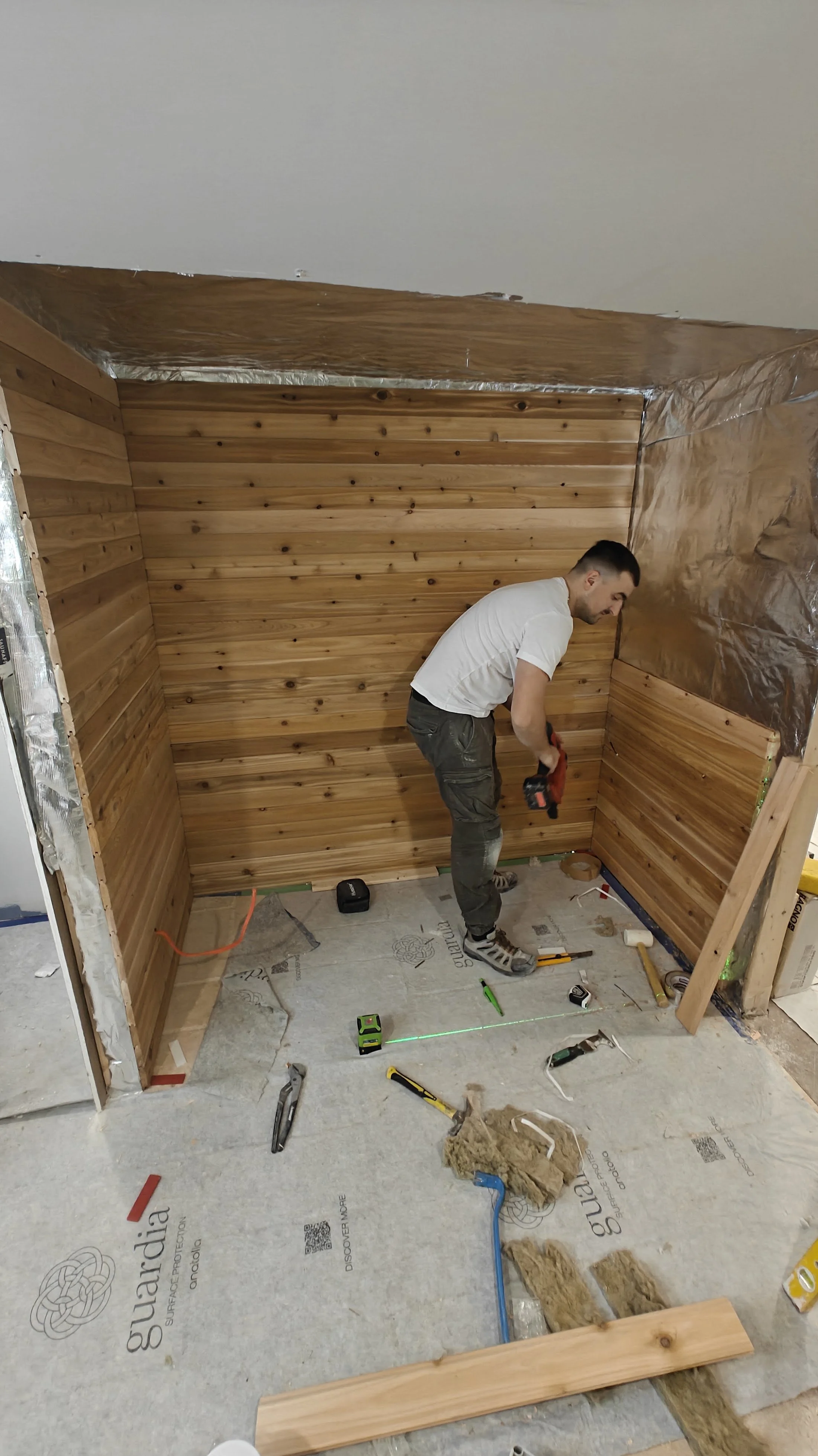 Man installing wooden paneling on a wall in a room under construction, with construction tools and insulation materials scattered on the floor.