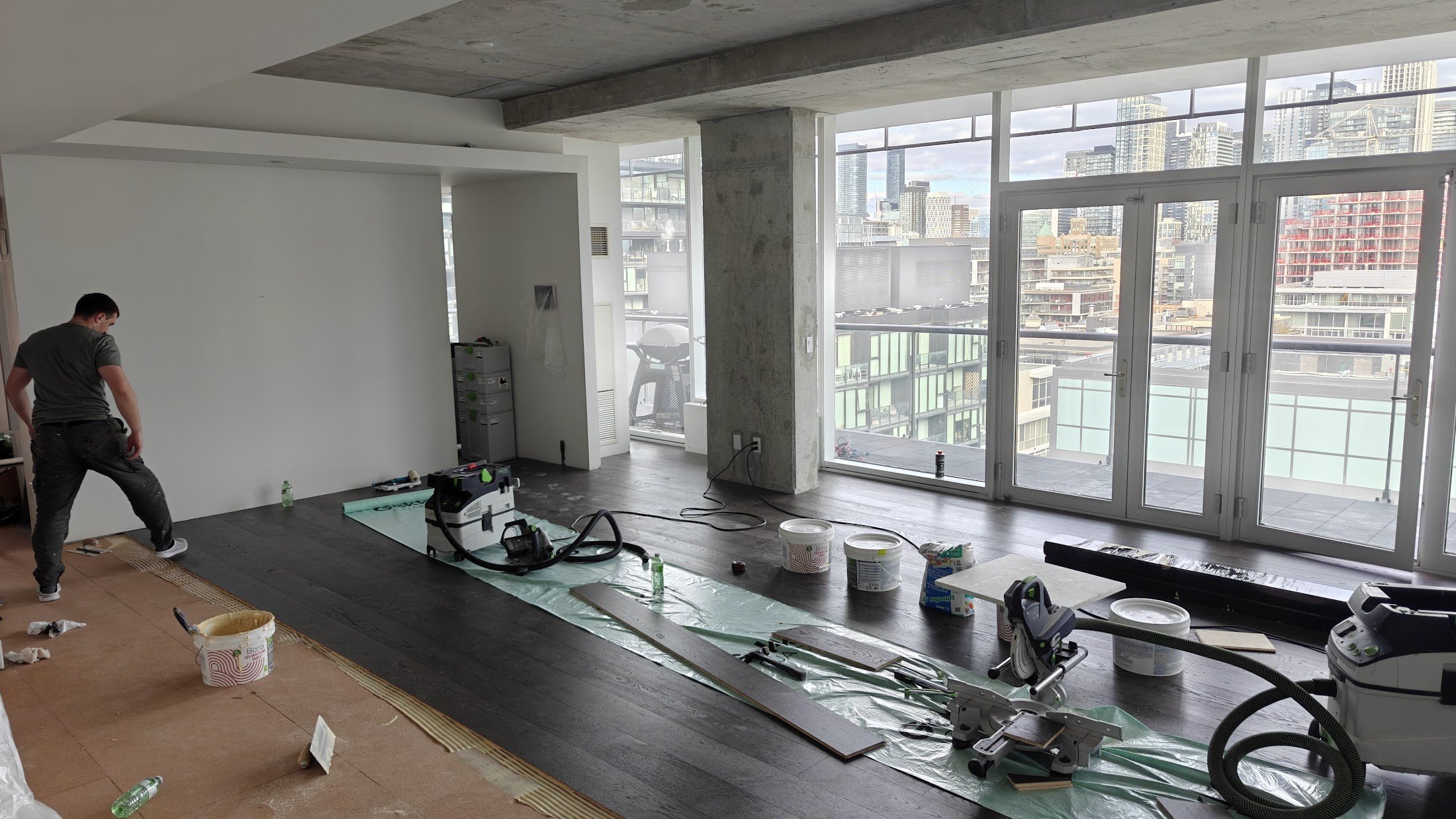 Interior of a high-rise apartment under renovation, with a worker installing dark hardwood flooring, large windows revealing a city skyline, and various renovation tools and supplies scattered around.