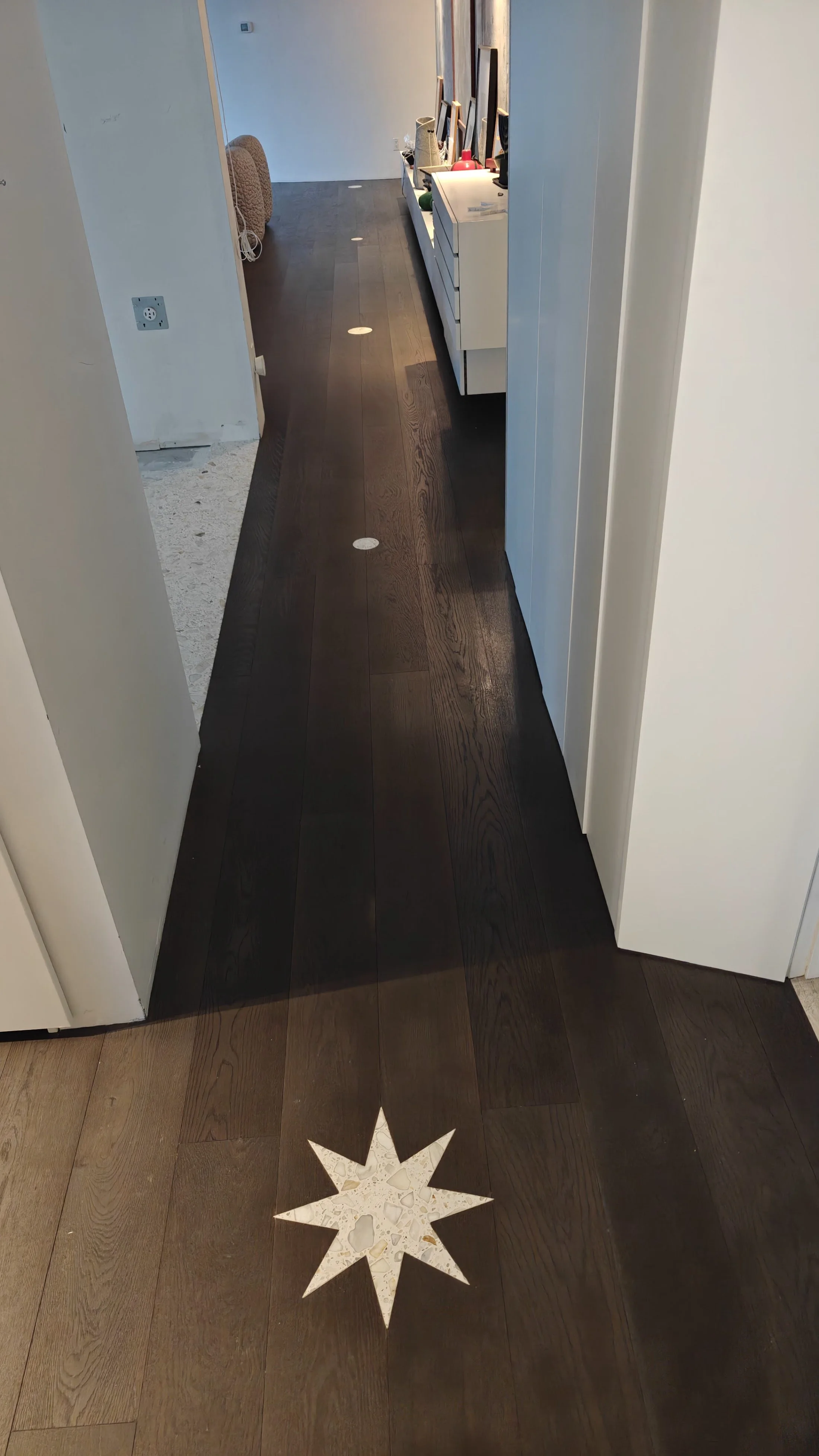 A hallway with dark wood flooring and a white star-shaped decoration on the floor. The hallway leads to a room with a white cabinet and various items on top, such as picture frames and decorative objects.