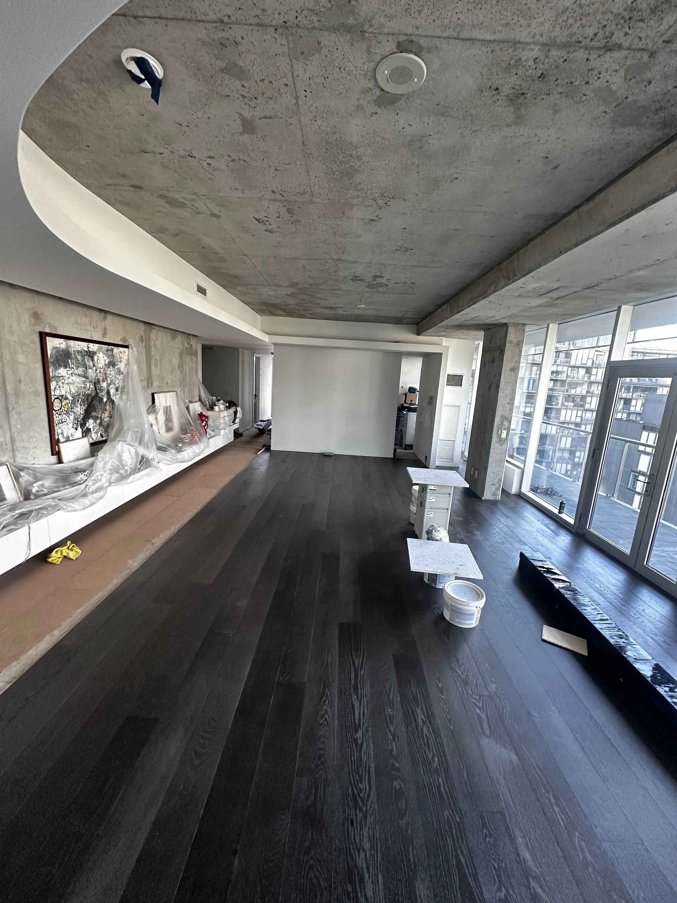 Modern living space under renovation with dark hardwood floors, large windows, various construction tools, and materials scattered on the floor.