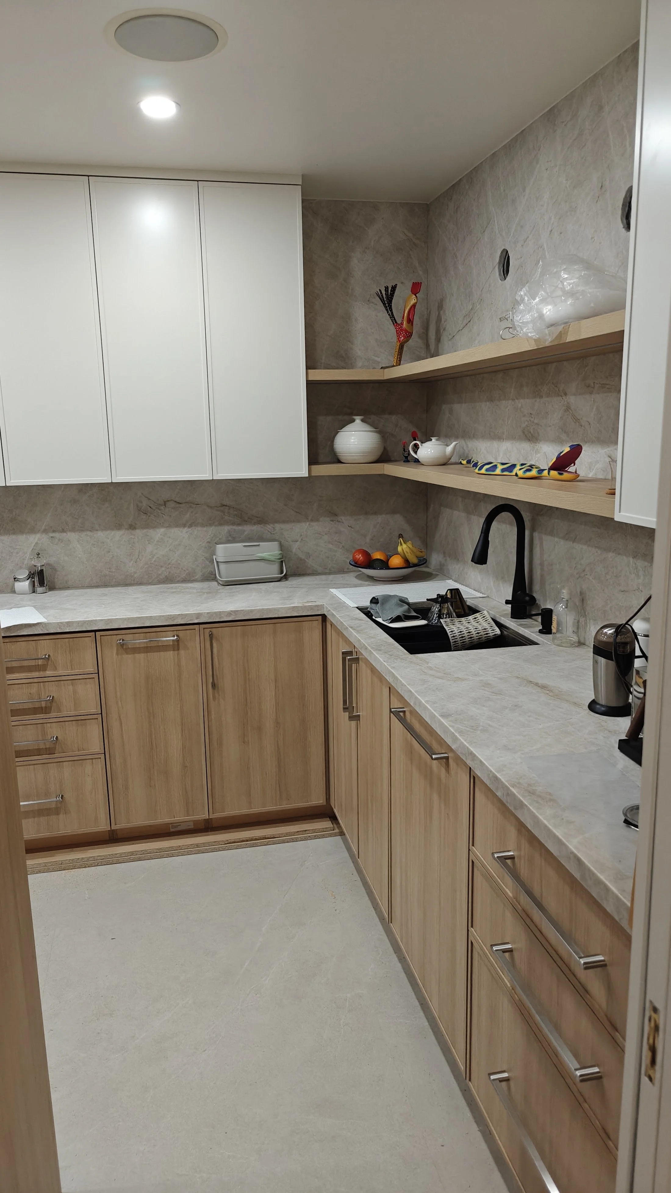 A modern kitchen with light wood cabinets, a beige marble countertop, and open wooden shelves. There is a black faucet over a sink, a bowl of fruit, and various kitchen utensils and appliances. White cabinets are on the wall above the countertop.