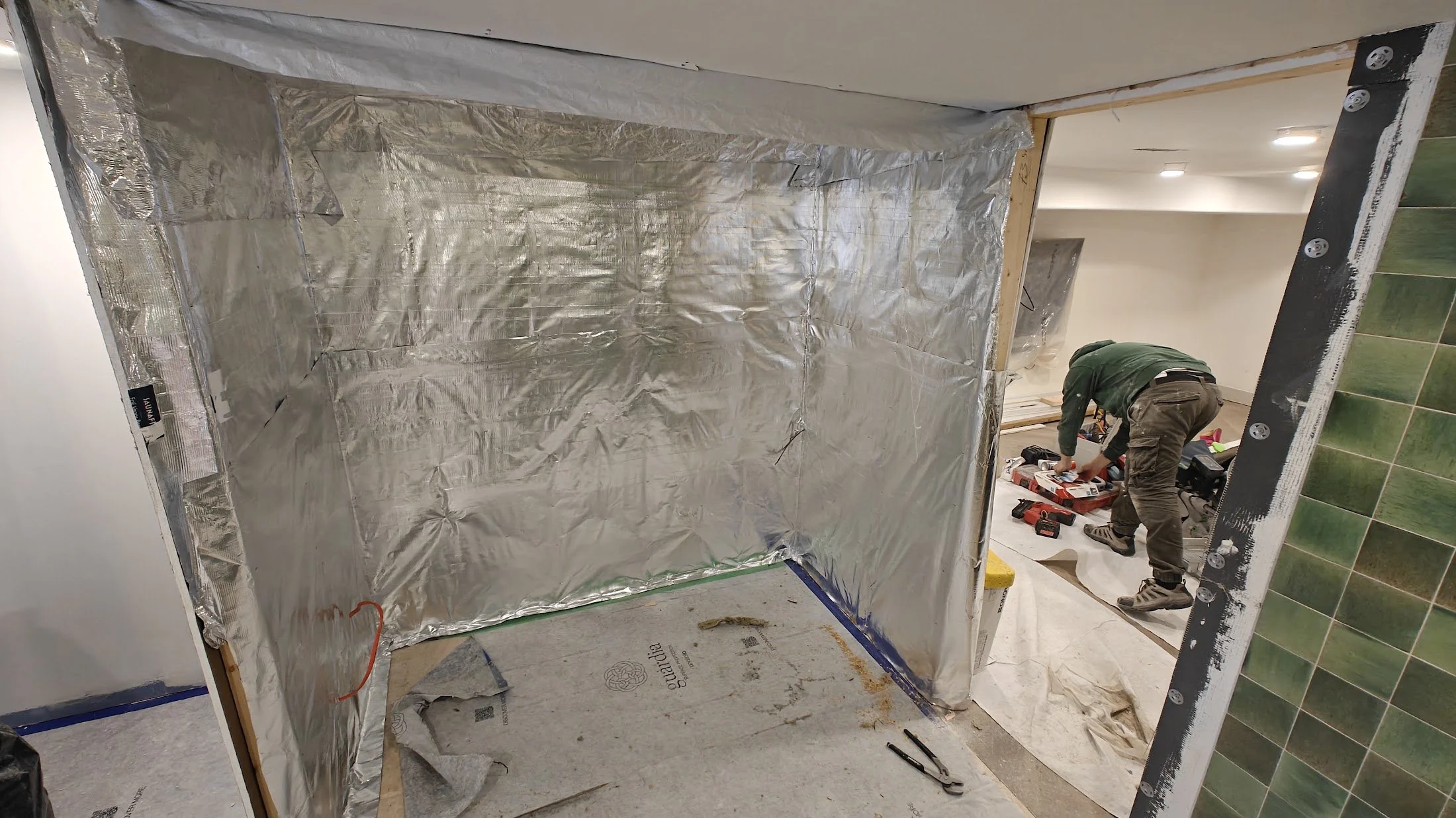 A person working on a construction project inside a room, installing or repairing tiles on the wall, with shiny insulation or vapor barrier material covering part of the space and green tiled wall visible on the right.