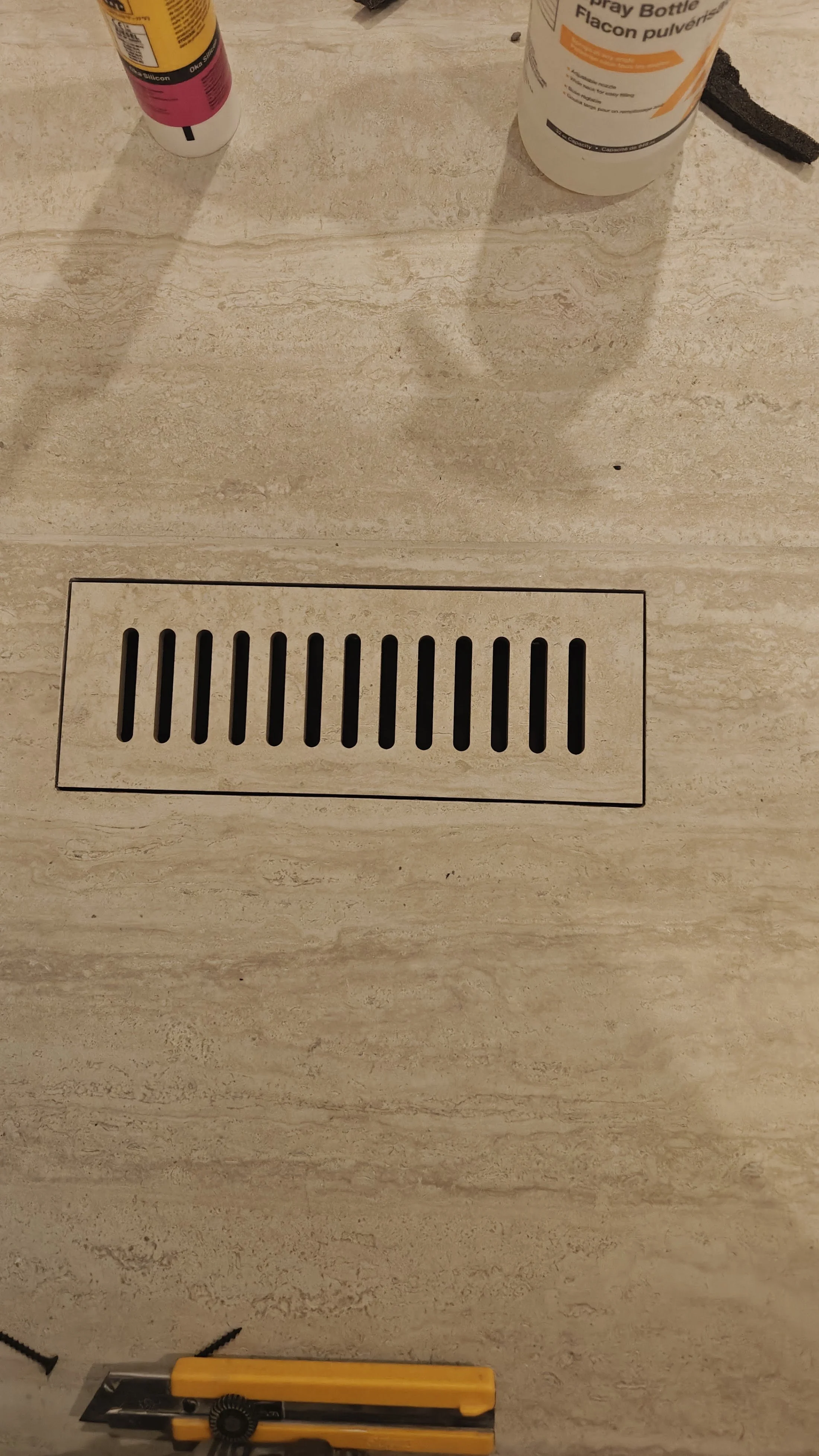 A floor drain with rectangular black slats in a beige tiled floor, partially covered by tools and bottles on the surface above.