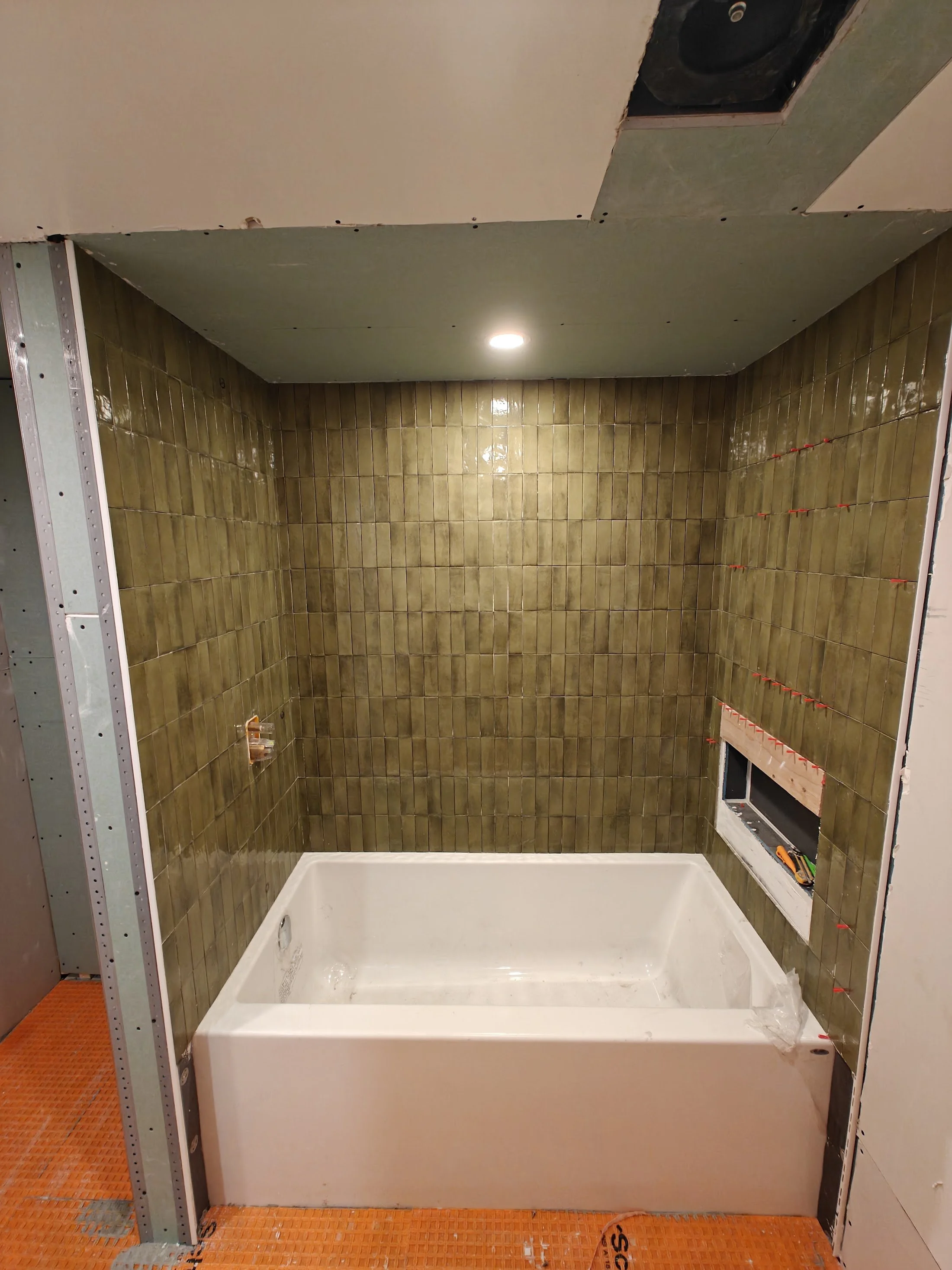 Bathroom under construction with tiled walls, a bathtub, and a ceiling with a recessed light, partially installed drywall, and a window opening.