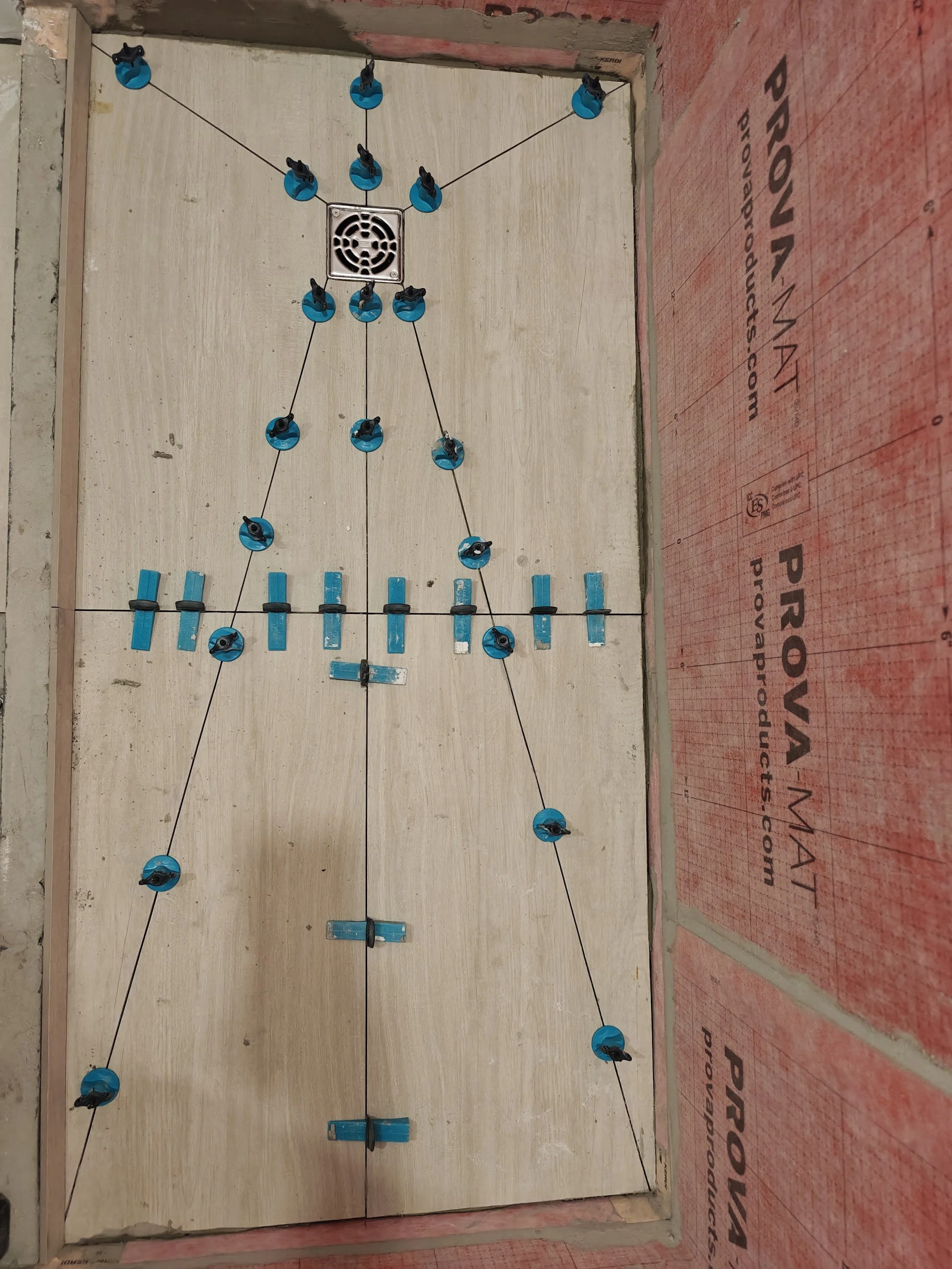 A partially constructed interior wall with a mounted steel drain cover at the top center. Blue plastic clips are attached to black strings arranged in a geometric pattern. There are some blue foam blocks and black clips along the wall at the bottom. 