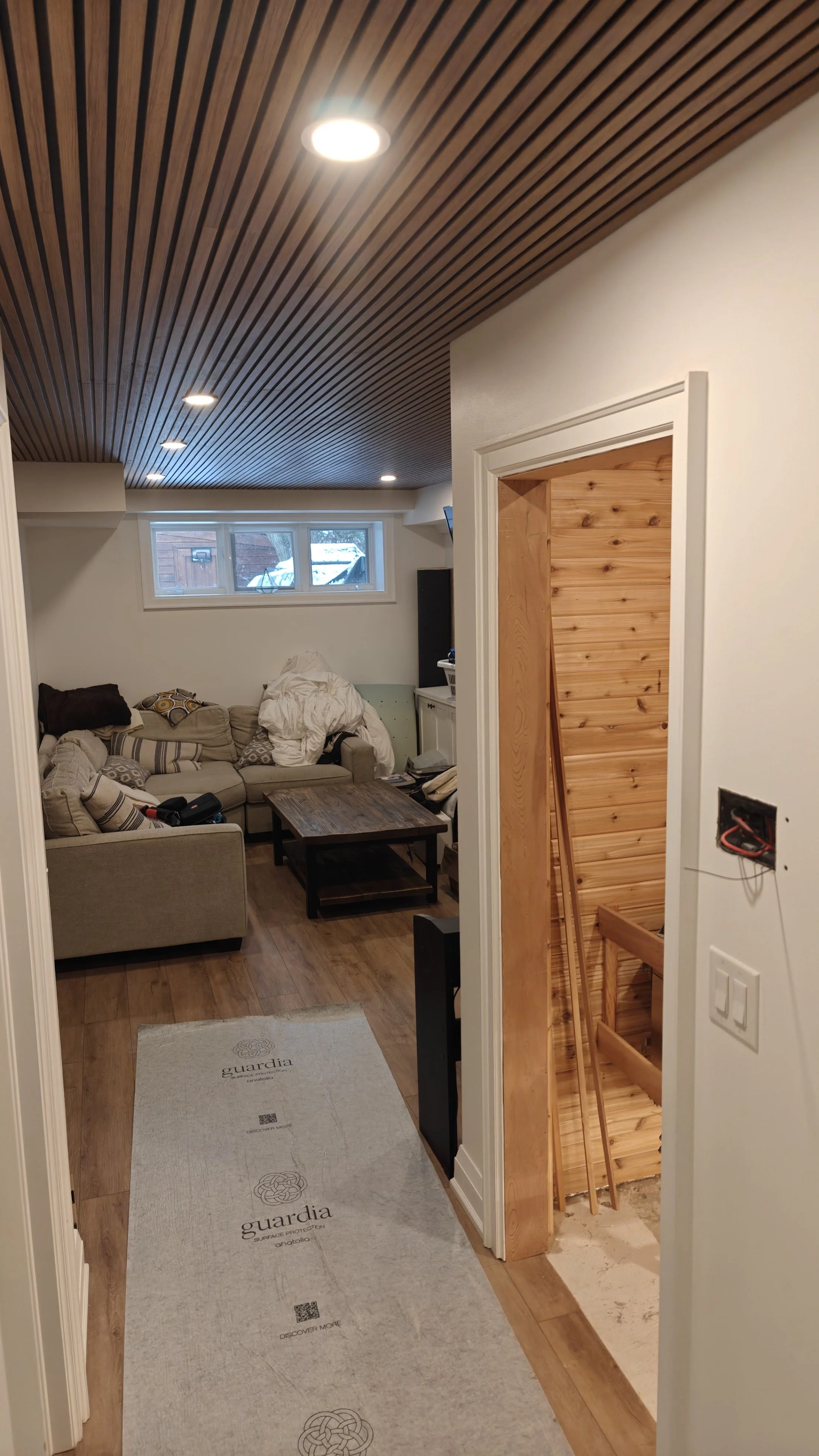 Living room with beige sofa, dark wood coffee table, and window showing outdoor snow. Wooden ceiling with recessed lights, hardwood floors, and unfinished staircase with wood paneling.