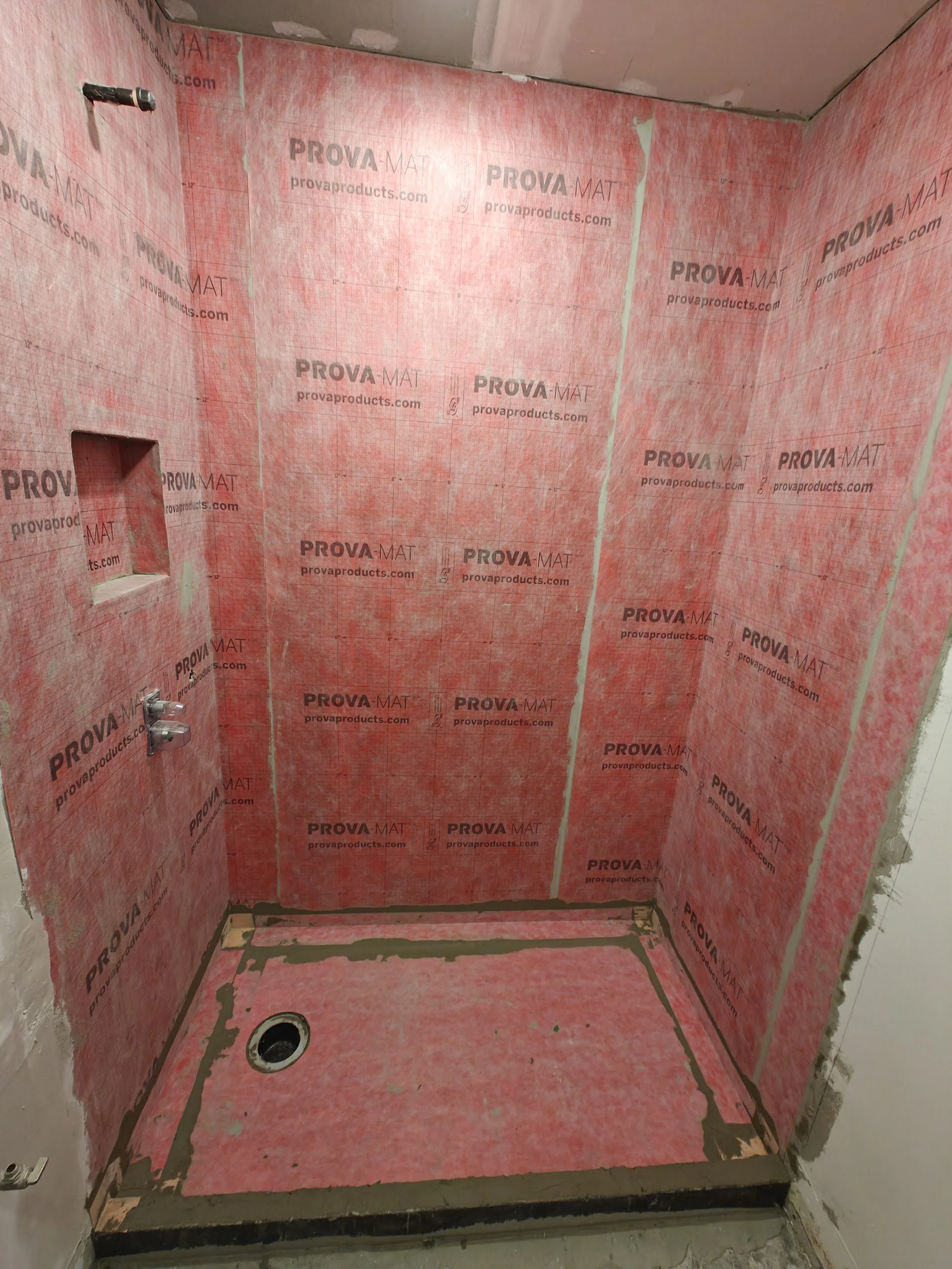 Bathroom shower stall under construction with pink waterproofing membrane on the walls and floor, a drain in the floor, and plumbing fixtures on the wall.