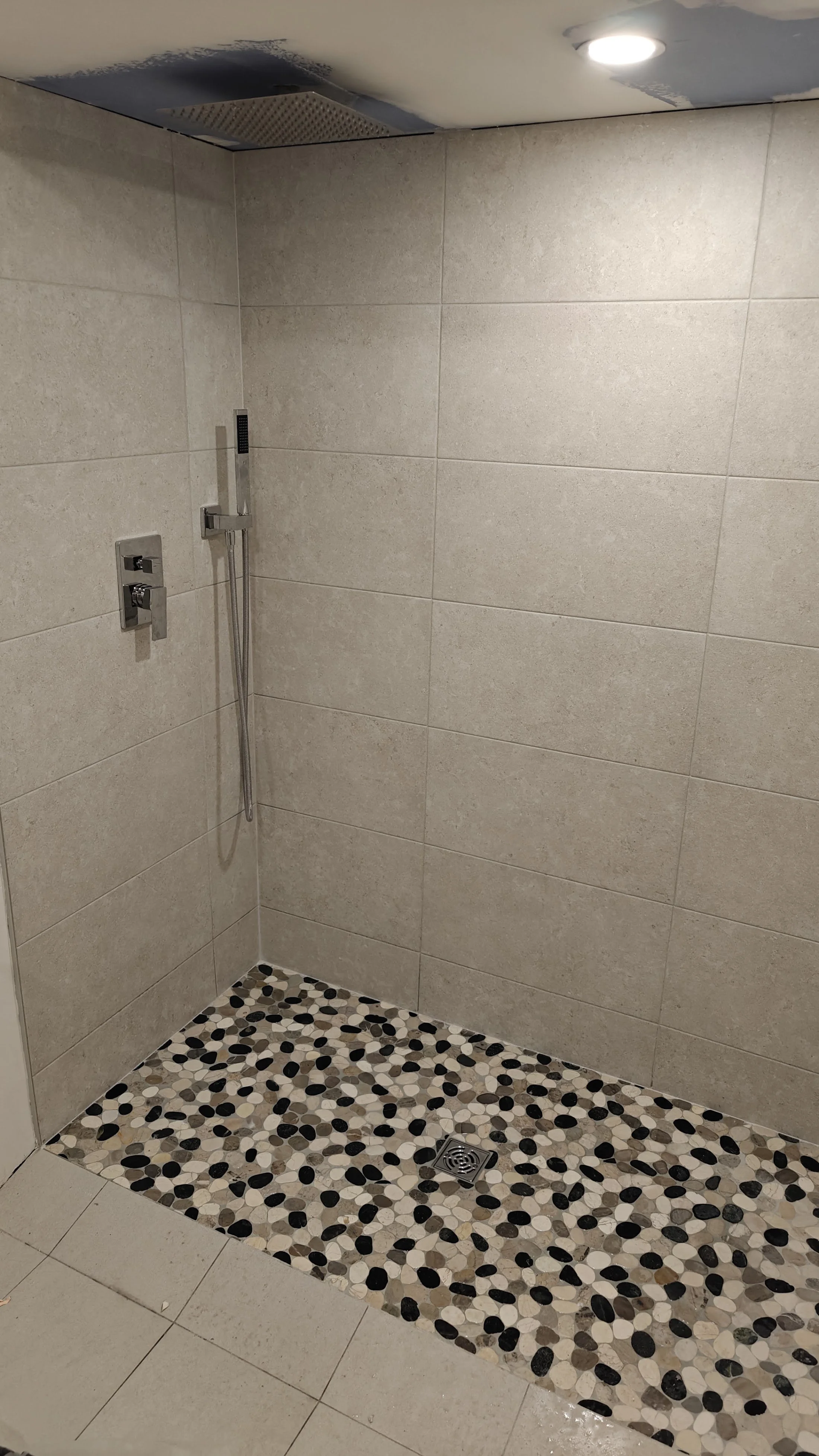 A beige tiled walk-in shower with a pebble floor section that has a silver drain. The shower has a modern chrome handle and adjustable shower head, with a vent on the ceiling and a recessed light. The walls are tiled with large rectangular beige tile