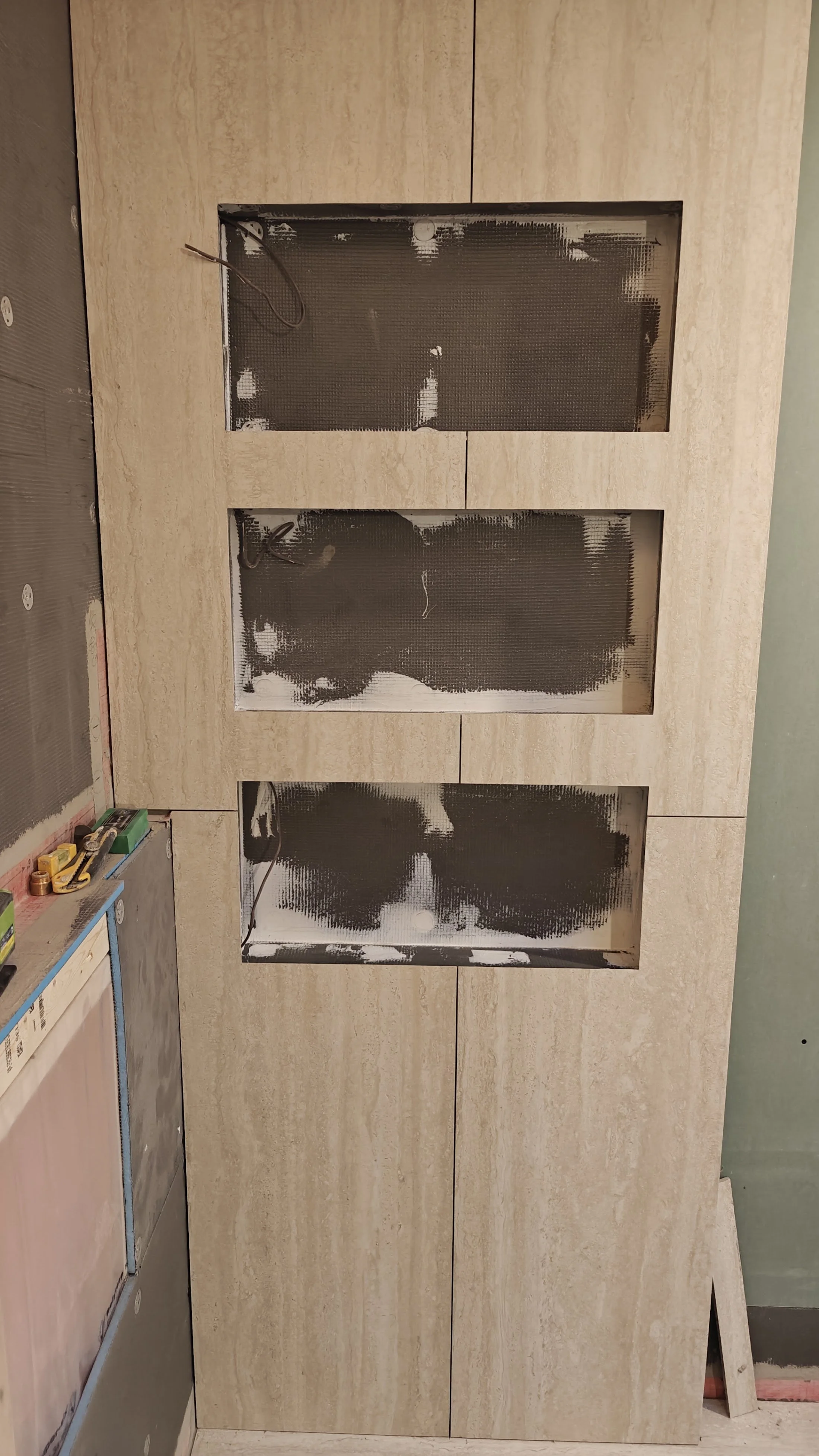 The image shows a partially installed wooden cabinet with three glass-front doors in a room under construction. Construction tools and supplies are visible on a small cart on the left side.