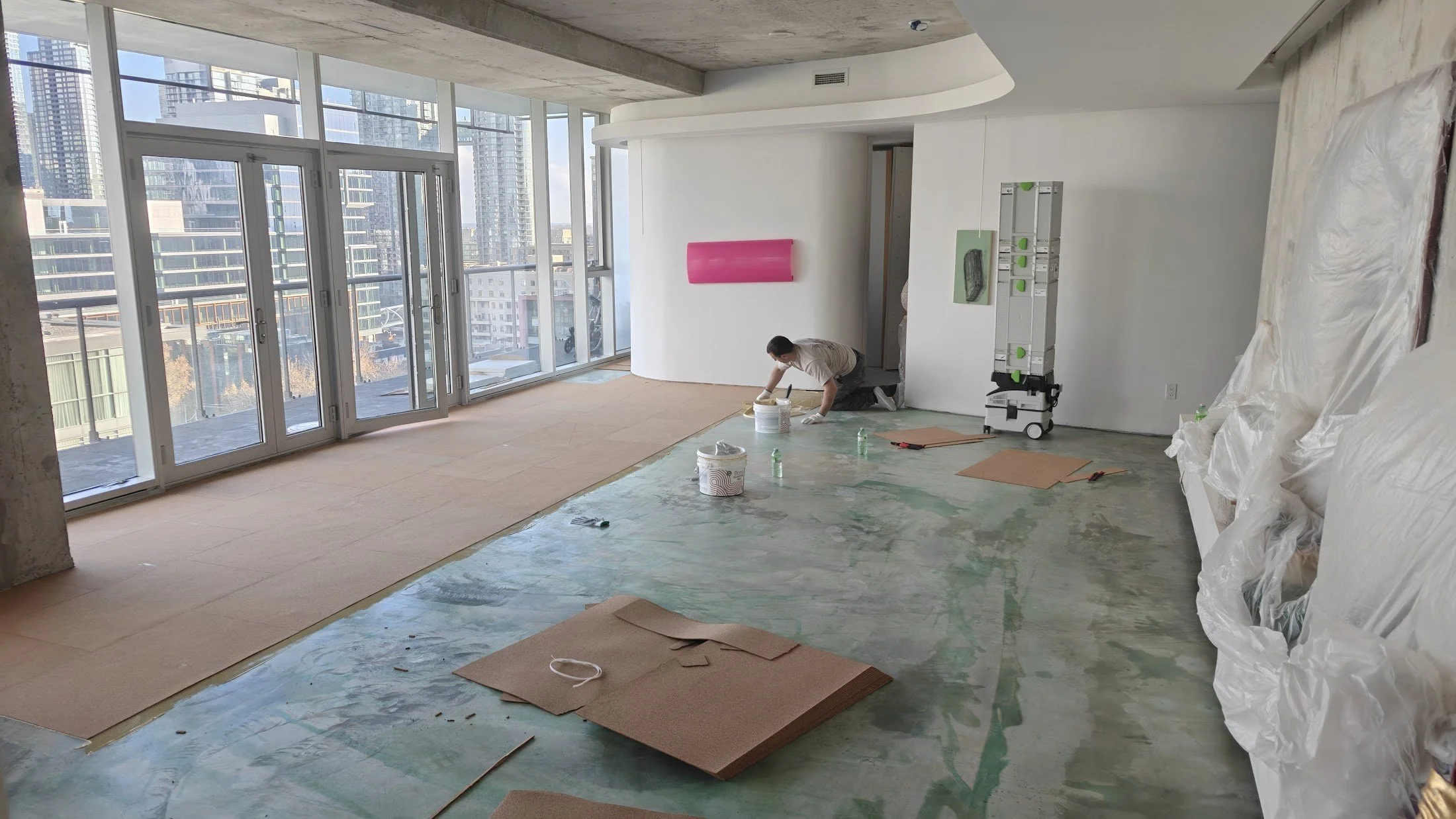 An interior space under renovation with large floor-to-ceiling windows showing a city skyline, a worker kneeling on the floor, surrounded by construction materials and tools, with partially installed flooring and plastic sheeting.