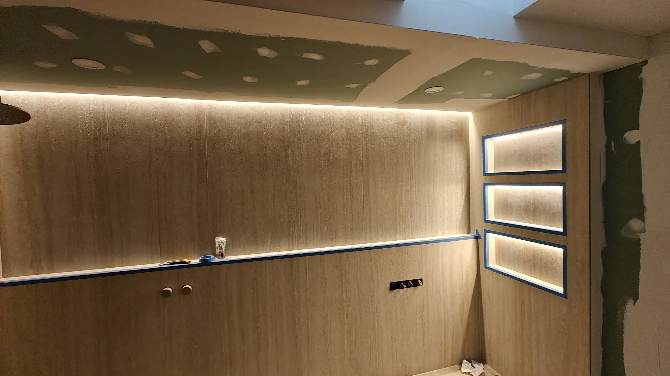 Partially constructed interior wall with framed openings, green drywall patches on ceiling, and recessed lighting. Tools and materials are on the floor and a small table.