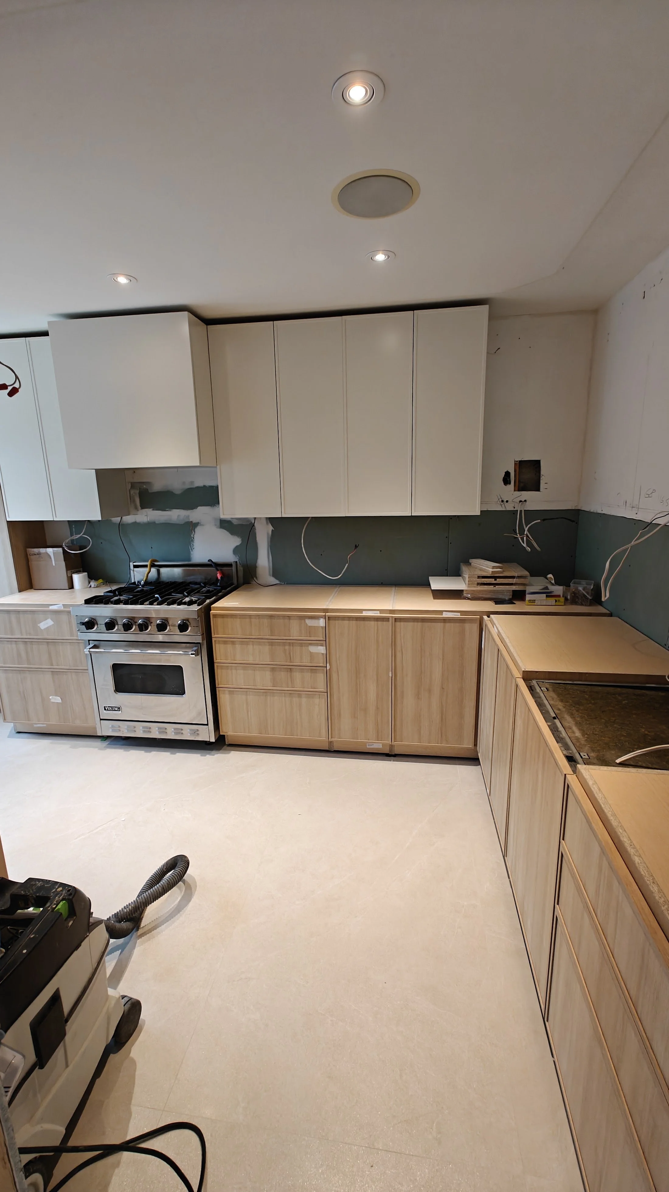 Kitchen under renovation with white upper cabinets, wooden lower cabinets, a stove, and exposed wiring, indicating ongoing work.