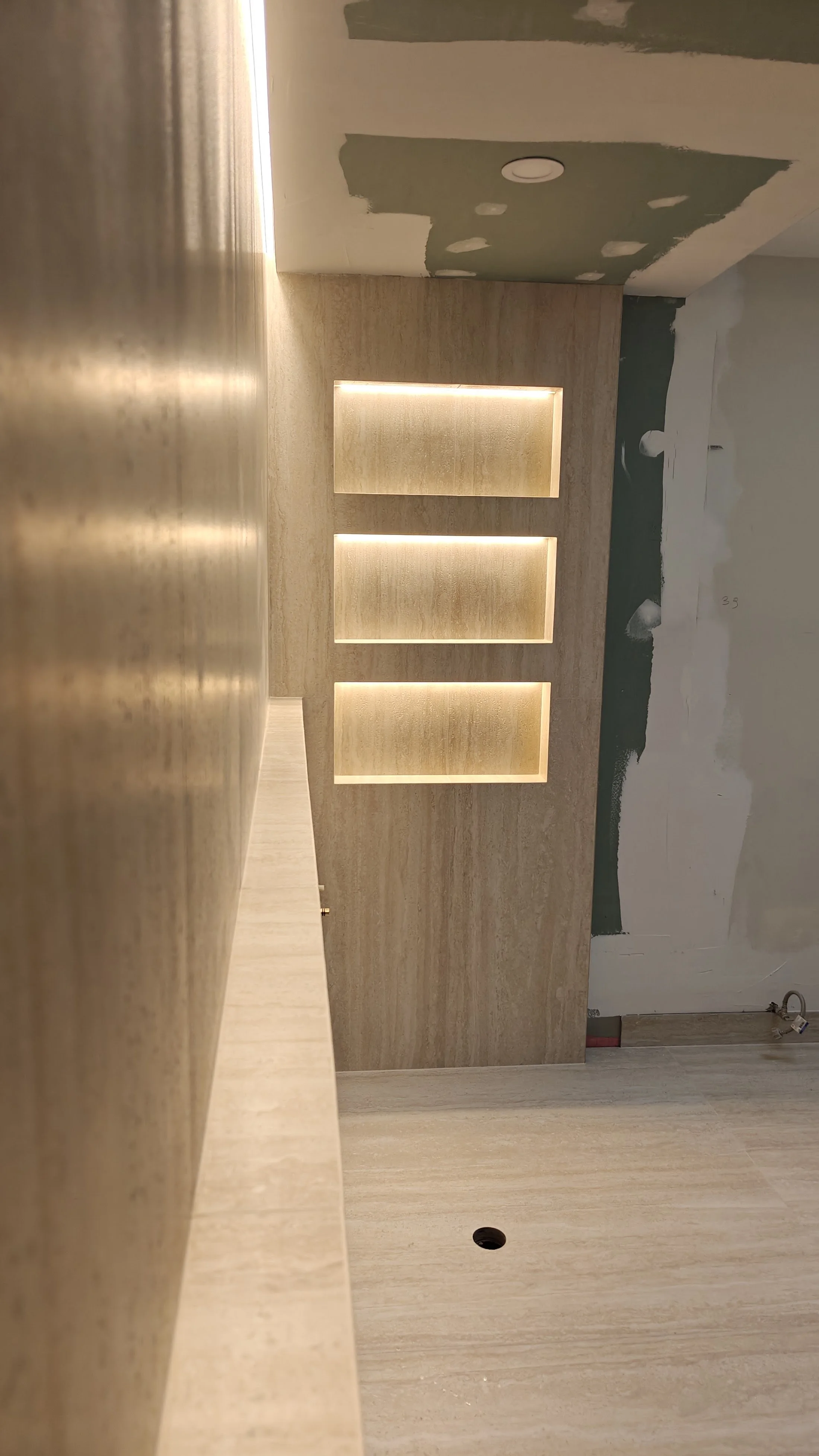 A partially finished room with a wooden wall featuring three backlit rectangular recesses, incomplete drywall with visible patches, and a beige countertop in the foreground.