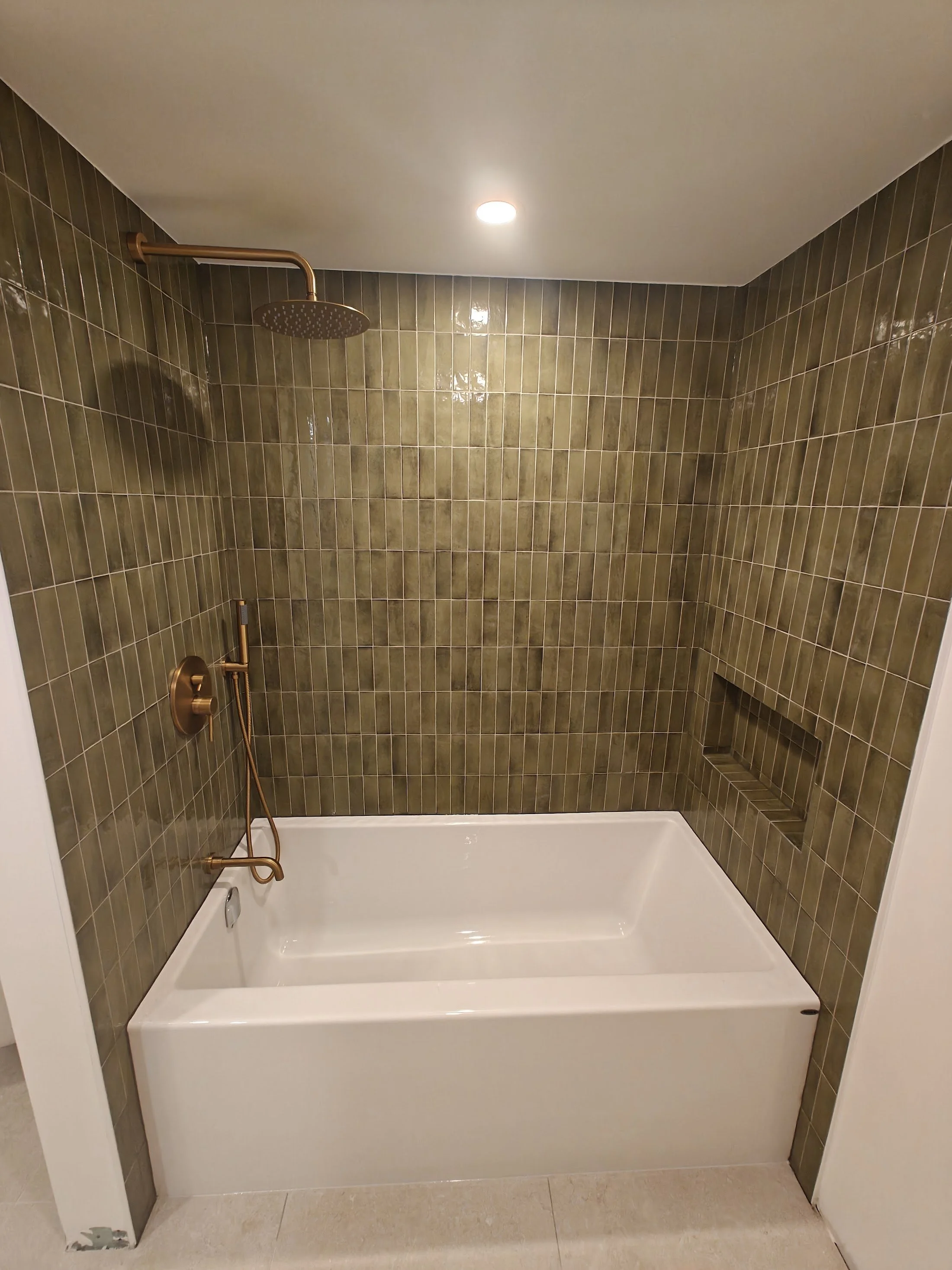 Bathroom shower with brown tiles, a gold showerhead, and a built-in shelf. The shower has a white bathtub and a beige tiled floor.