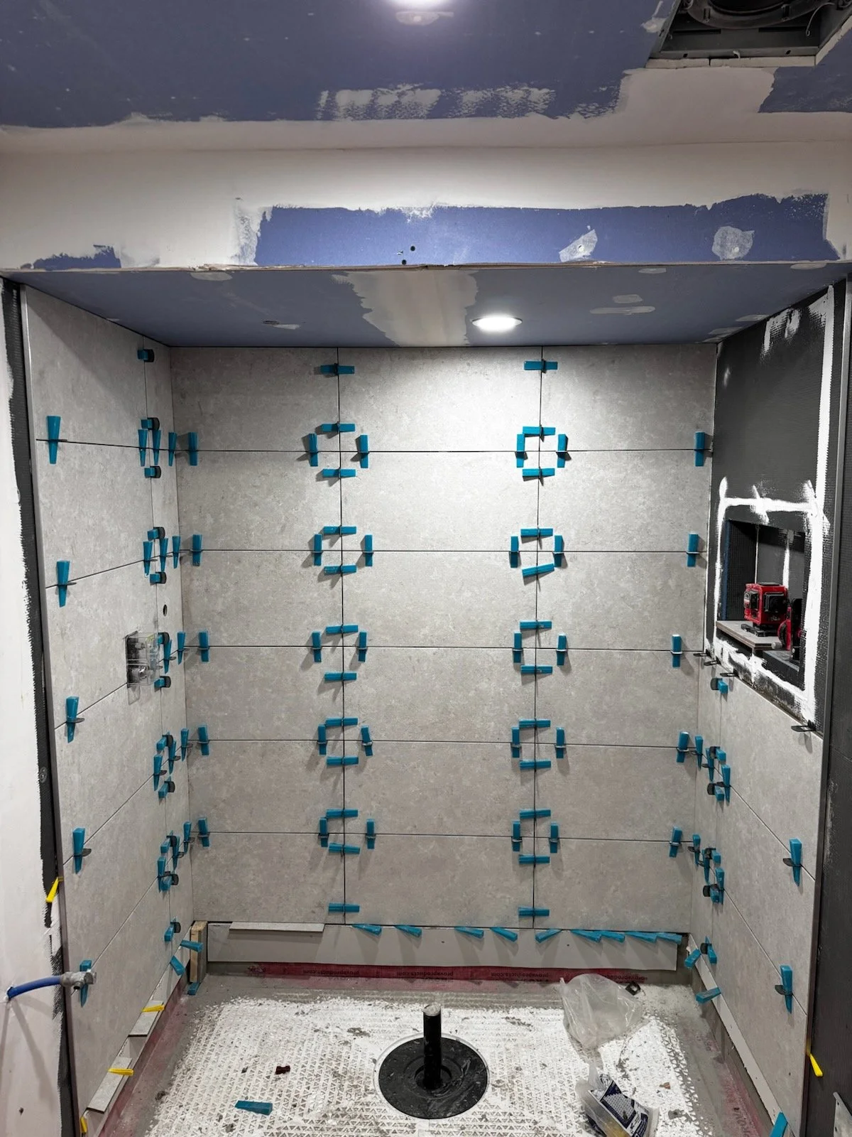 Bathroom under renovation with drywall tiles being installed, blue spacers are used to set the tiles evenly, a small toolbox and construction tools sit on the right wall, a circular fixture on the ceiling, and a hole for plumbing in the floor.