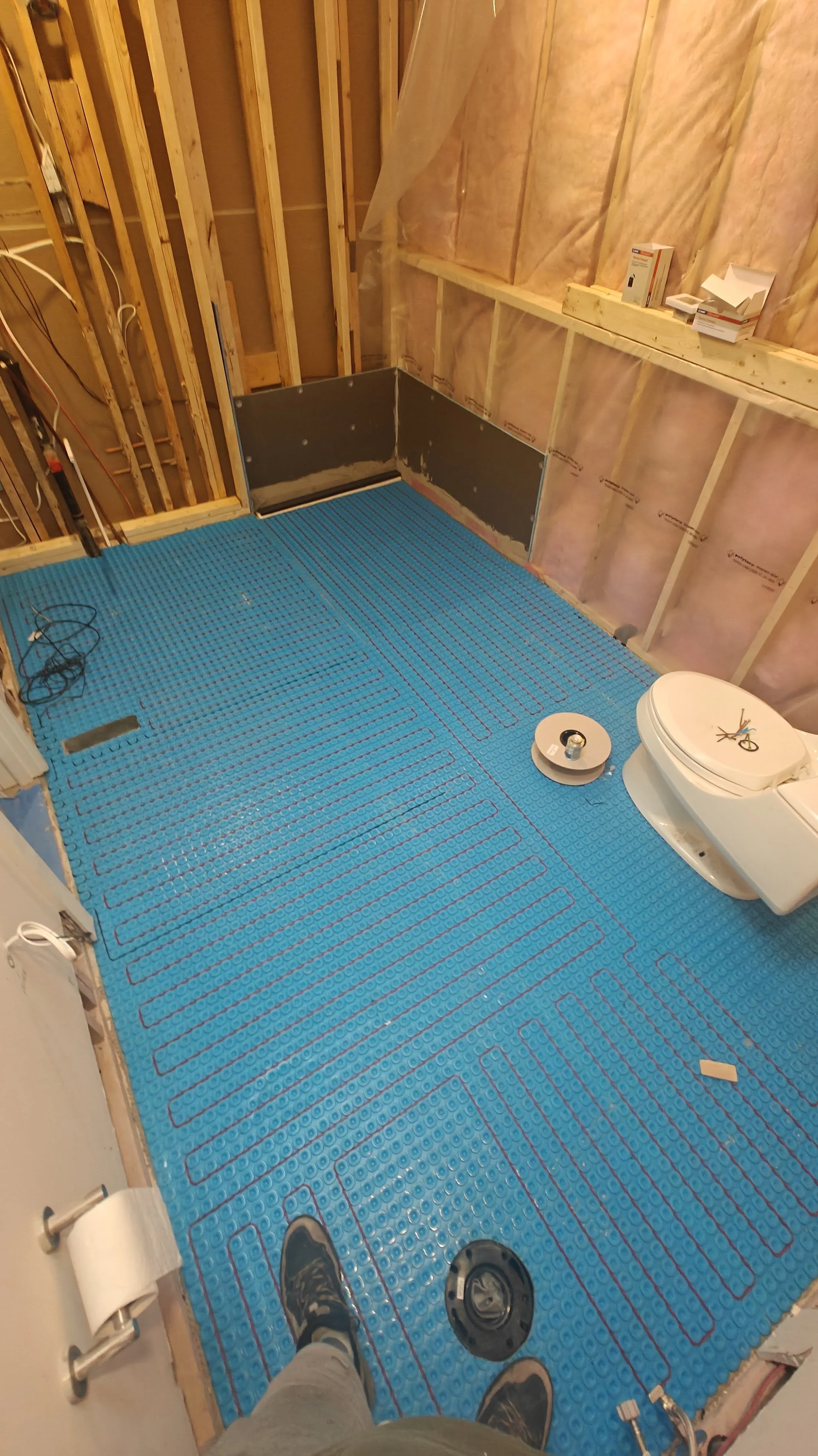 Bathroom under construction with blue electric radiant floor heating mats installed, toilet and tools visible, unfinished walls with insulation, and person in work boots taking the photo.
