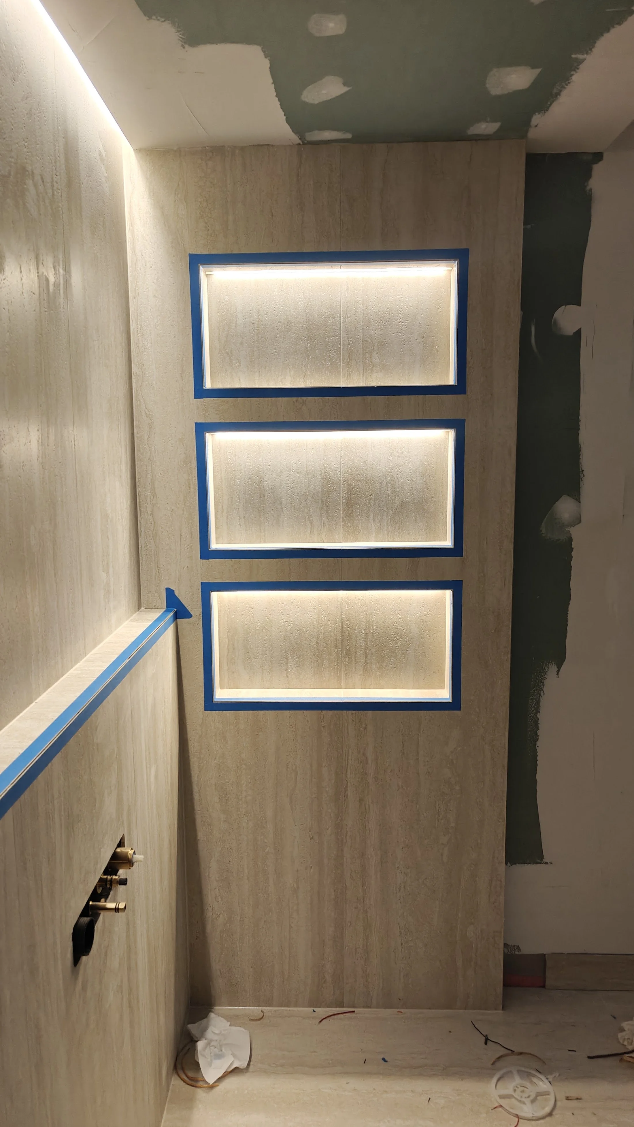 This image shows a room with three rectangular recessed lights on a beige wall, framed with blue tape. The ceiling is partially unfinished, with visible drywall patches and paint. The floor appears to be under construction, with tools, wiring, and de