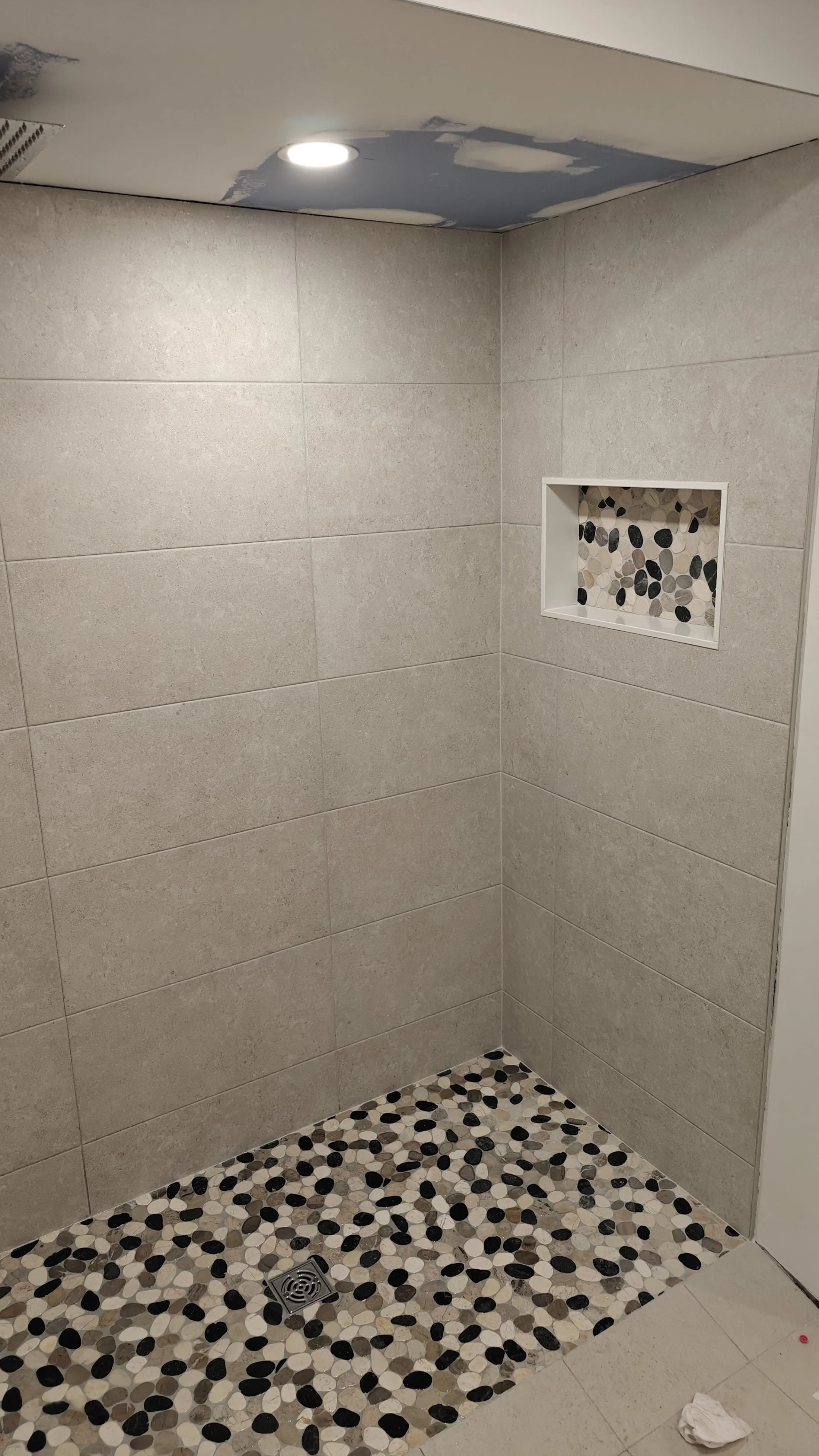 A corner of a shower with a beige tiled wall, a recessed niche with pebble tile, a pebble tiled shower floor, a ceiling with a recessed light, and a drain on the floor. Partially visible white cloth on the lower right.