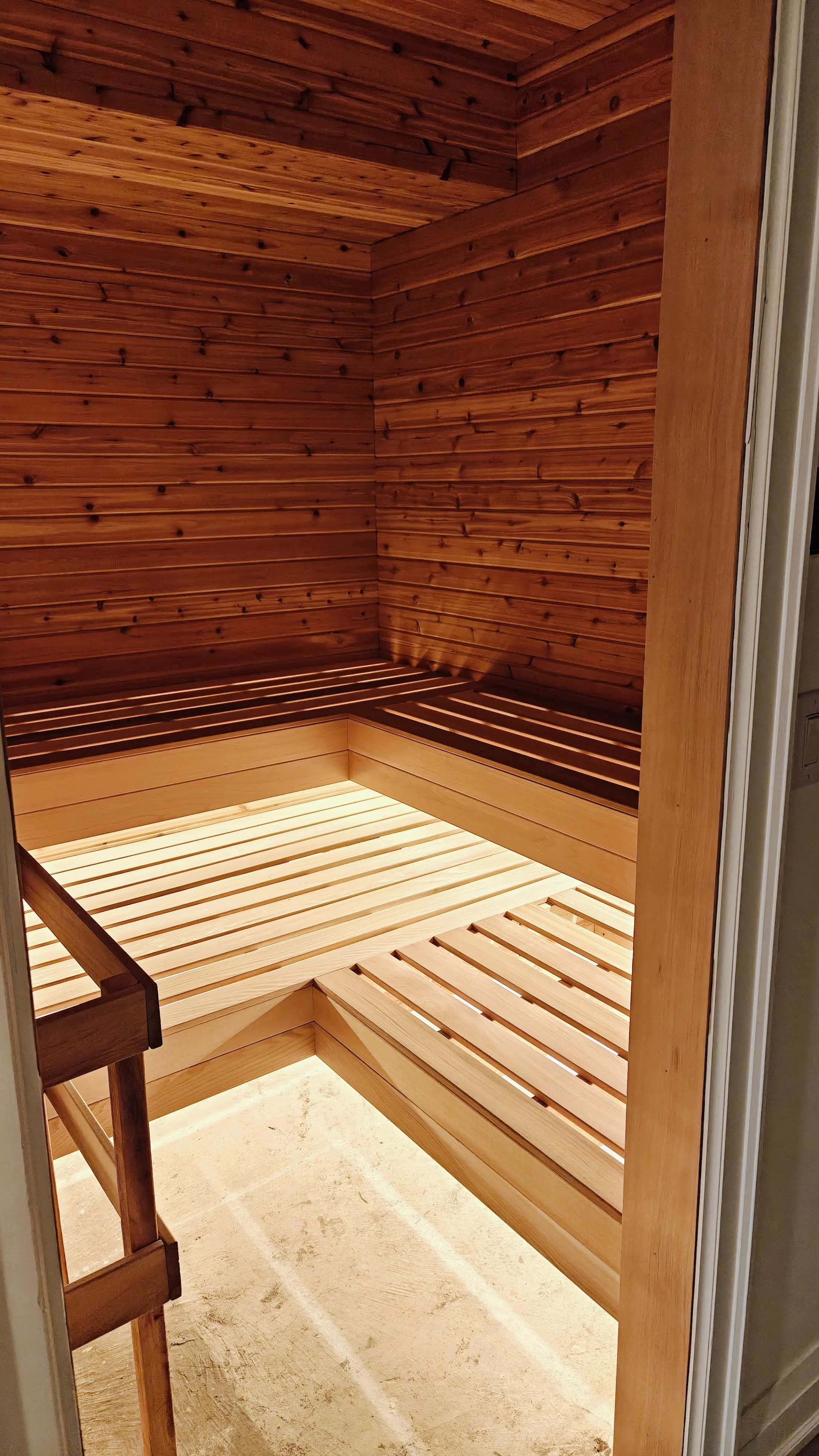 A small wooden room, possibly a sauna, with wooden slats on the walls, ceiling, and bench seating; the floor is a light-colored concrete or stone.