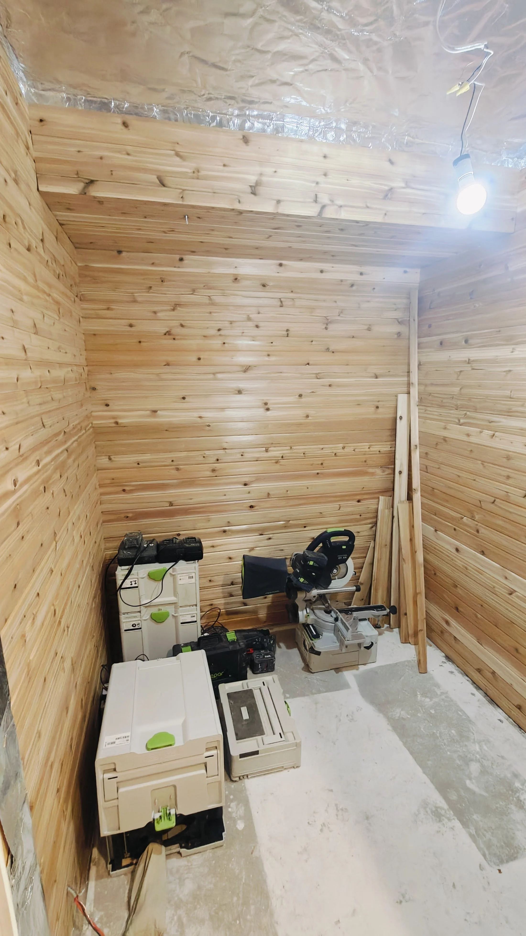 A small construction room with plywood walls and a ceiling, with construction tools and equipment including a miter saw, a cordless drill, and electrical boxes placed on the floor.