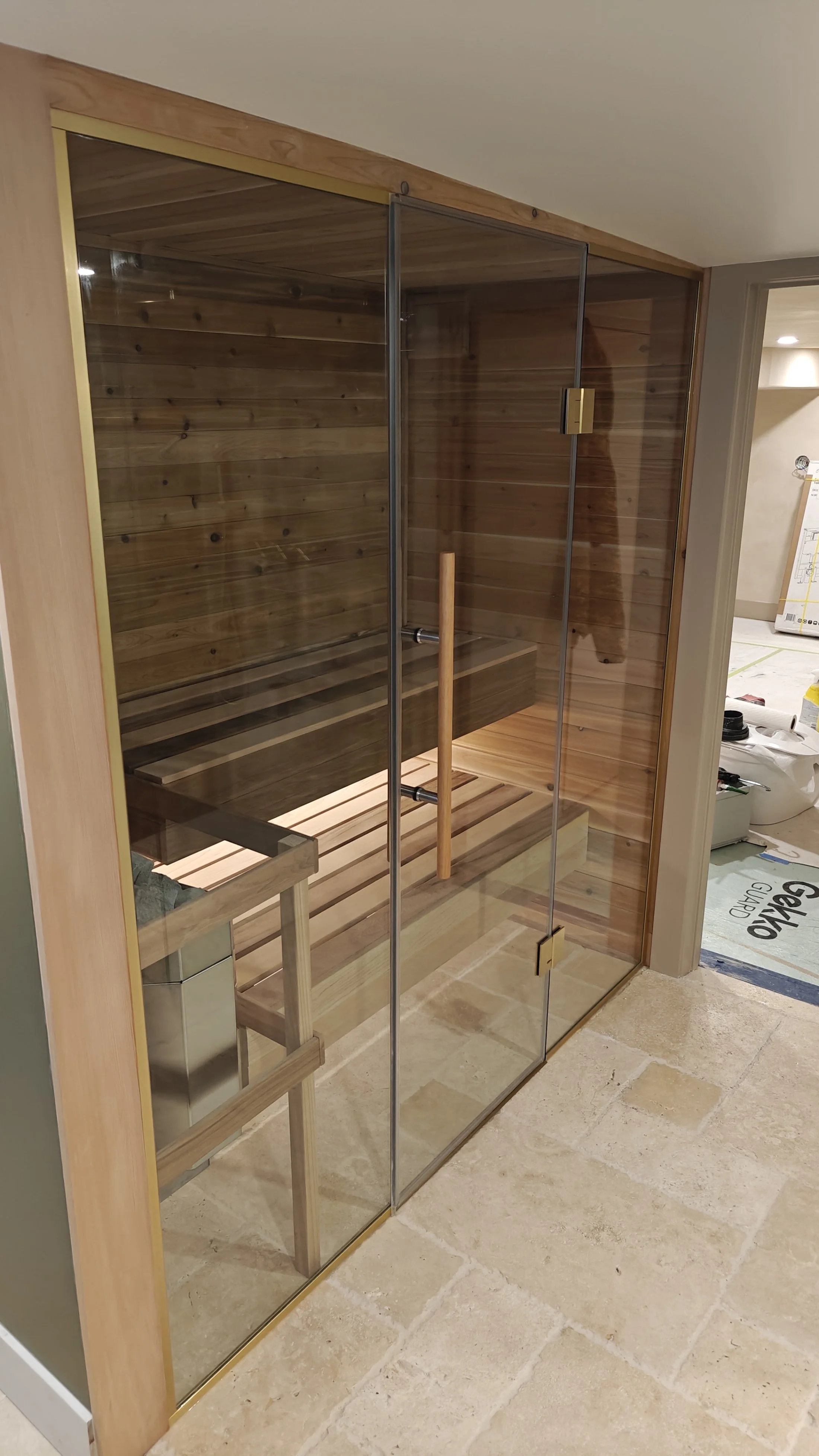 A glass-front sauna with wooden interior and seating, located in an indoor space. Construction supplies and unfinished flooring are visible nearby.