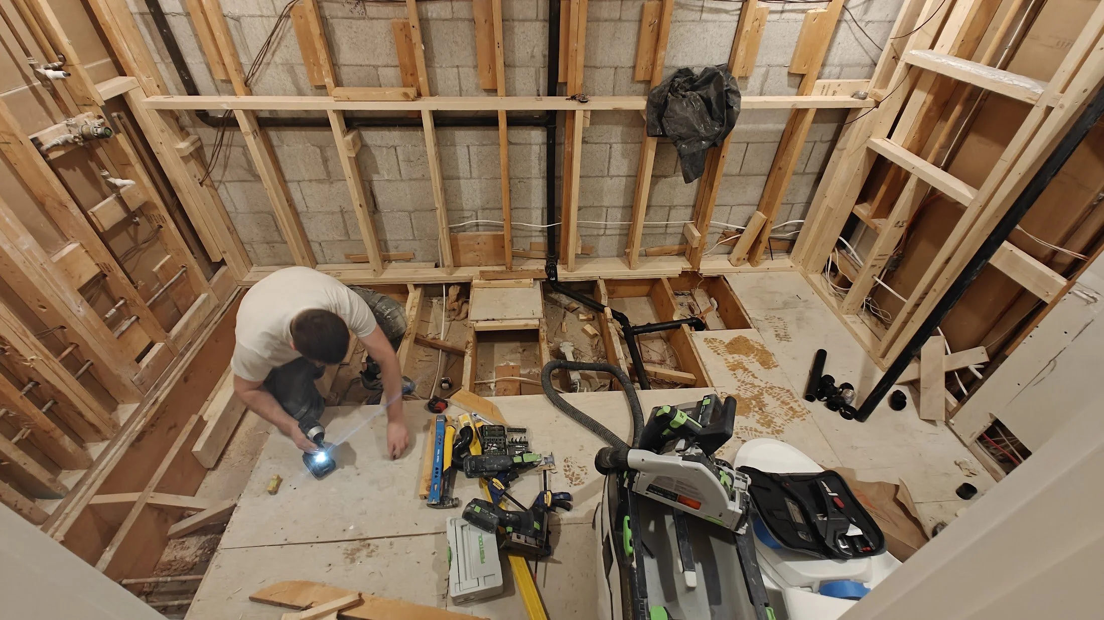 A person working on a home renovation project in a partially built room, surrounded by tools and construction materials, with exposed wood framing and a cinder block wall in the background.