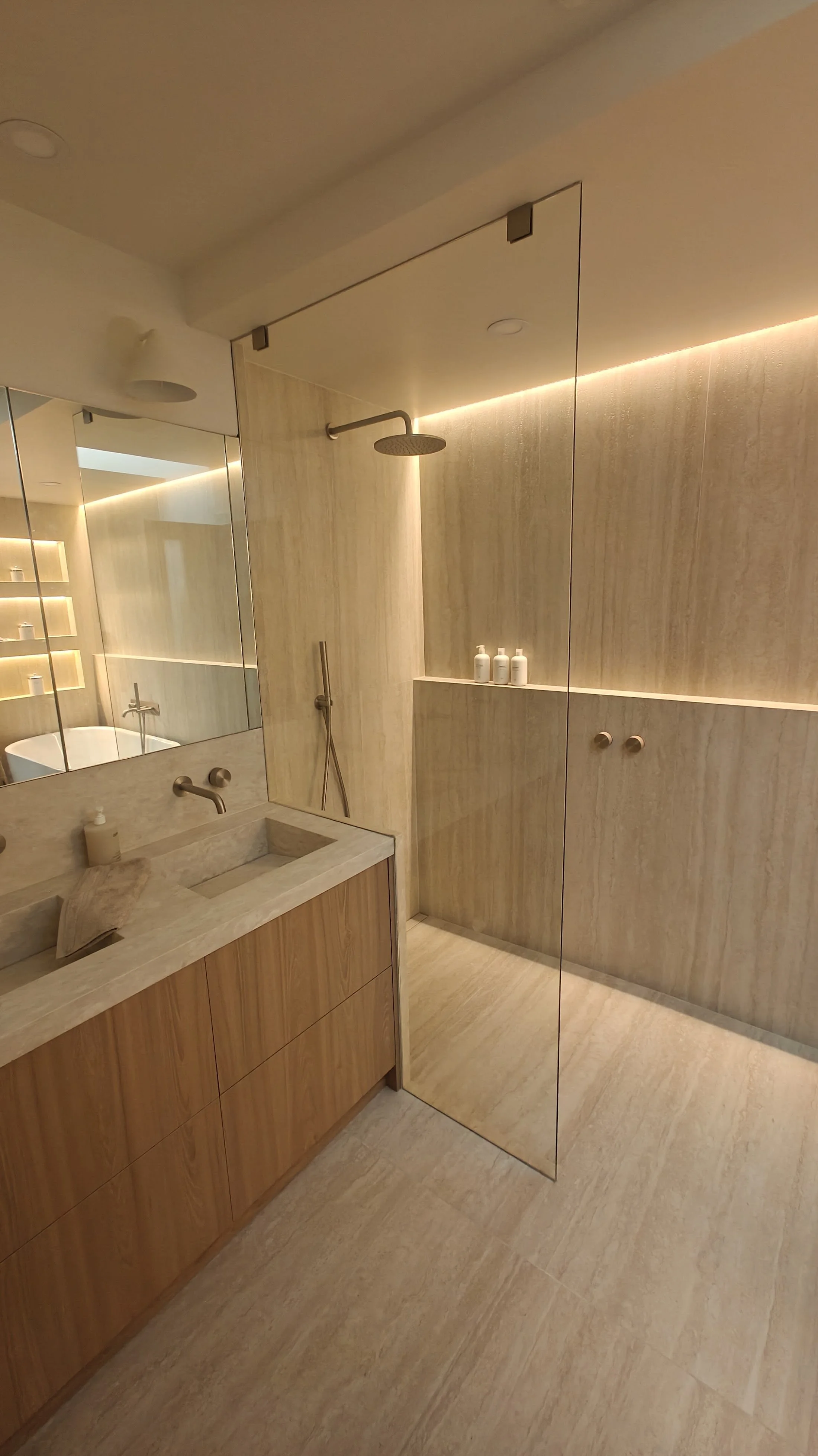 Modern bathroom with beige marble walls, a vanity with a built-in sink, a large mirror, and a separate shower area with a rainfall showerhead, illuminated by warm lighting.