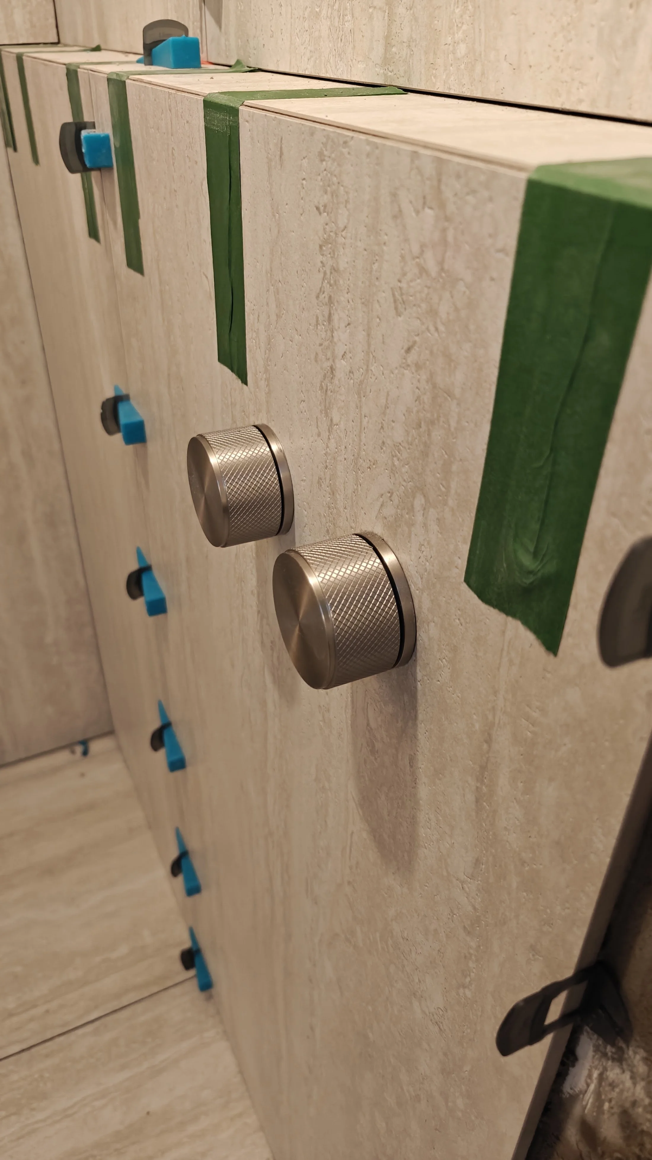 Close-up of a beige marble kitchen island with two silver knobs, a strip of green painter's tape and blue painter's tape tabs on the side, and a set of black and gray handles in the background.
