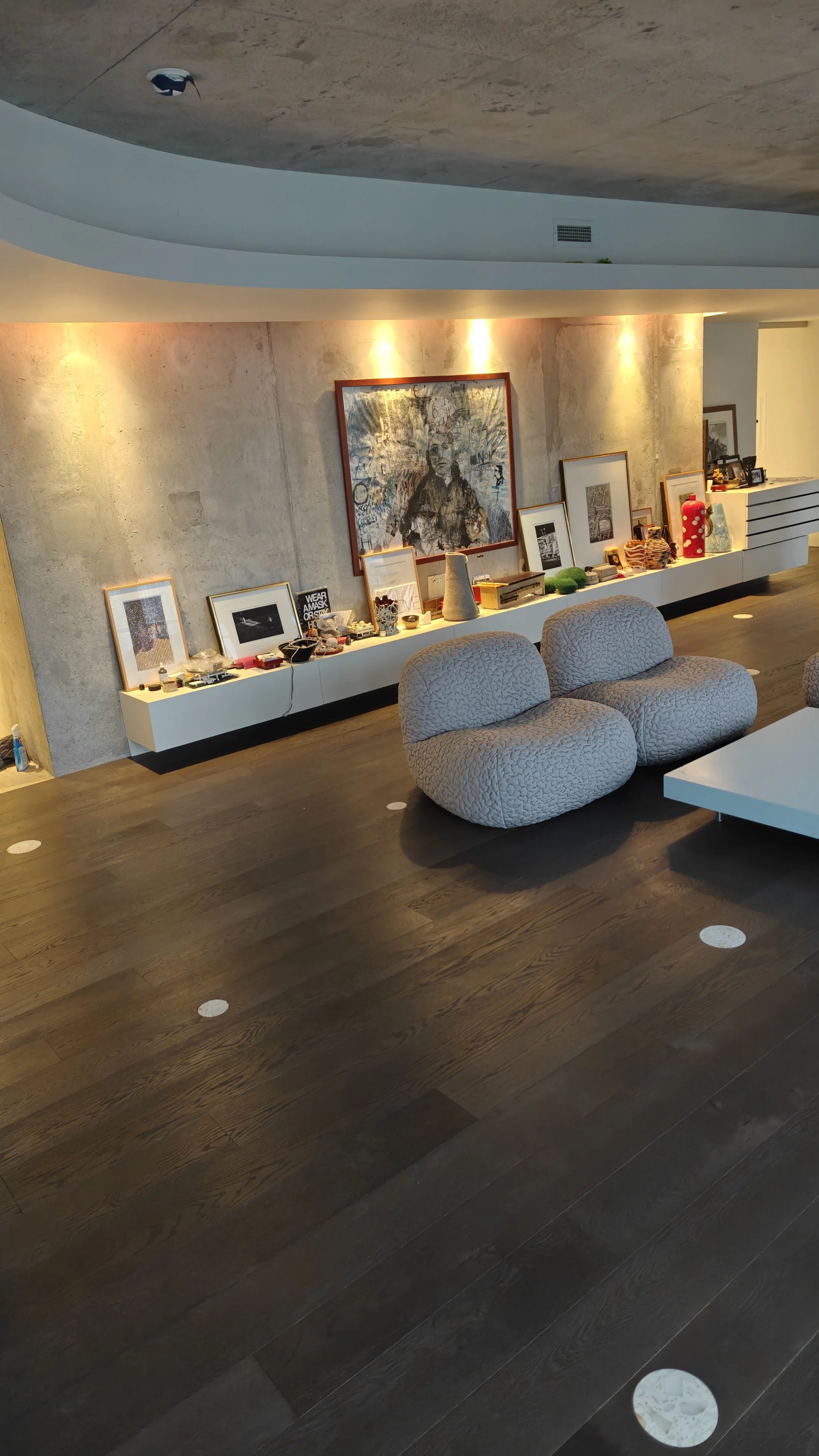 Living room with wooden flooring, modern rounded armchair, concrete wall with artwork and framed pictures, decorative objects on a white wall-mounted shelf, and a white ceiling with recessed lighting.
