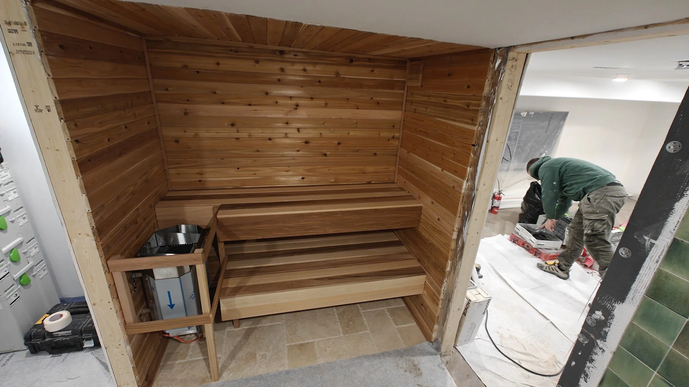 Interior wooden sauna with bentwood benches and wood-paneled walls under construction, with a worker installing or working on the setup.