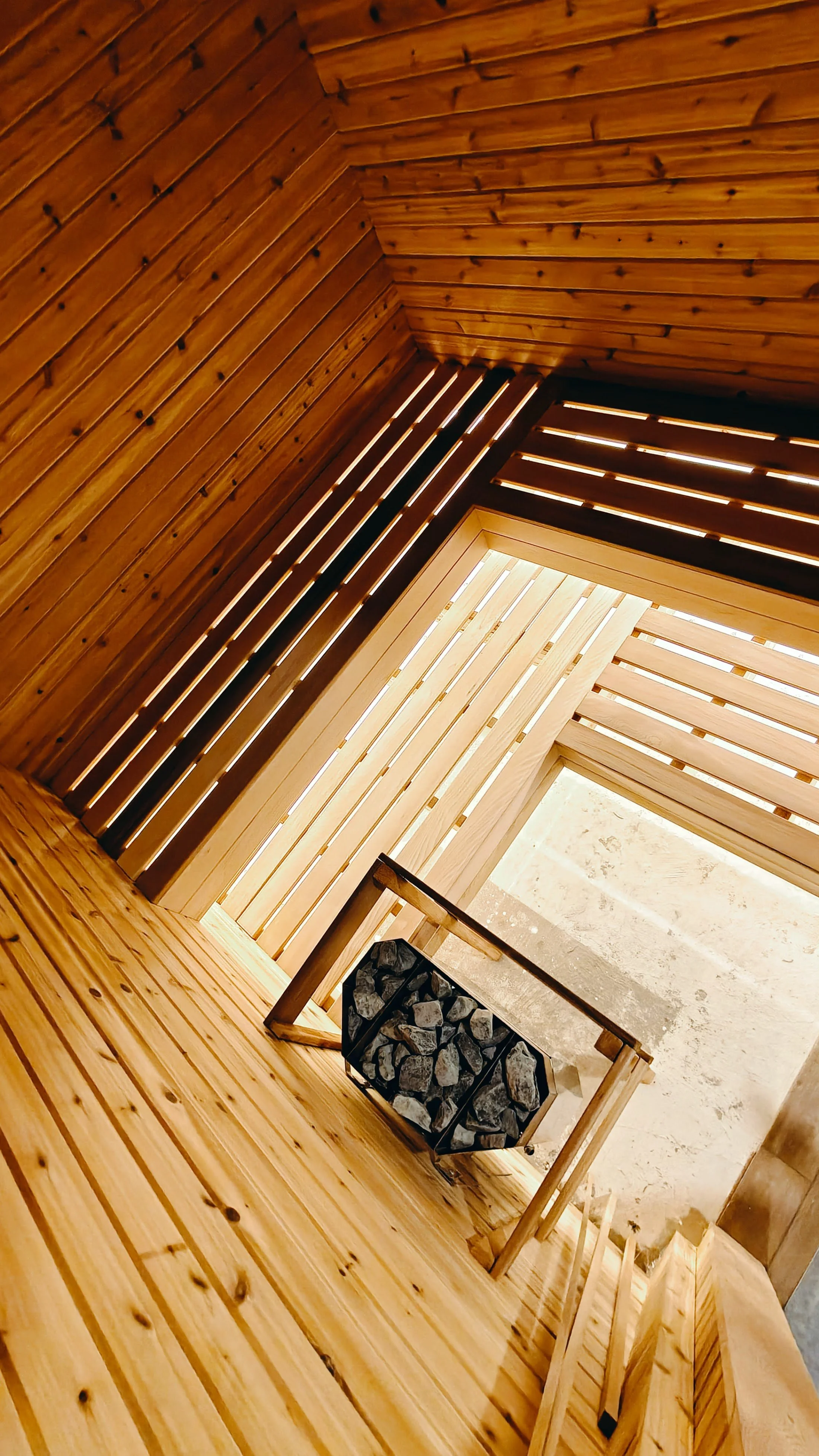 Interior view of a wooden cabin with walls, ceiling, and floor made of pine wood planks. A small chair with a stone-themed cushion is near a framed fireplace or wood storage area. Horizontal wooden slats cover the window area, allowing some natural l