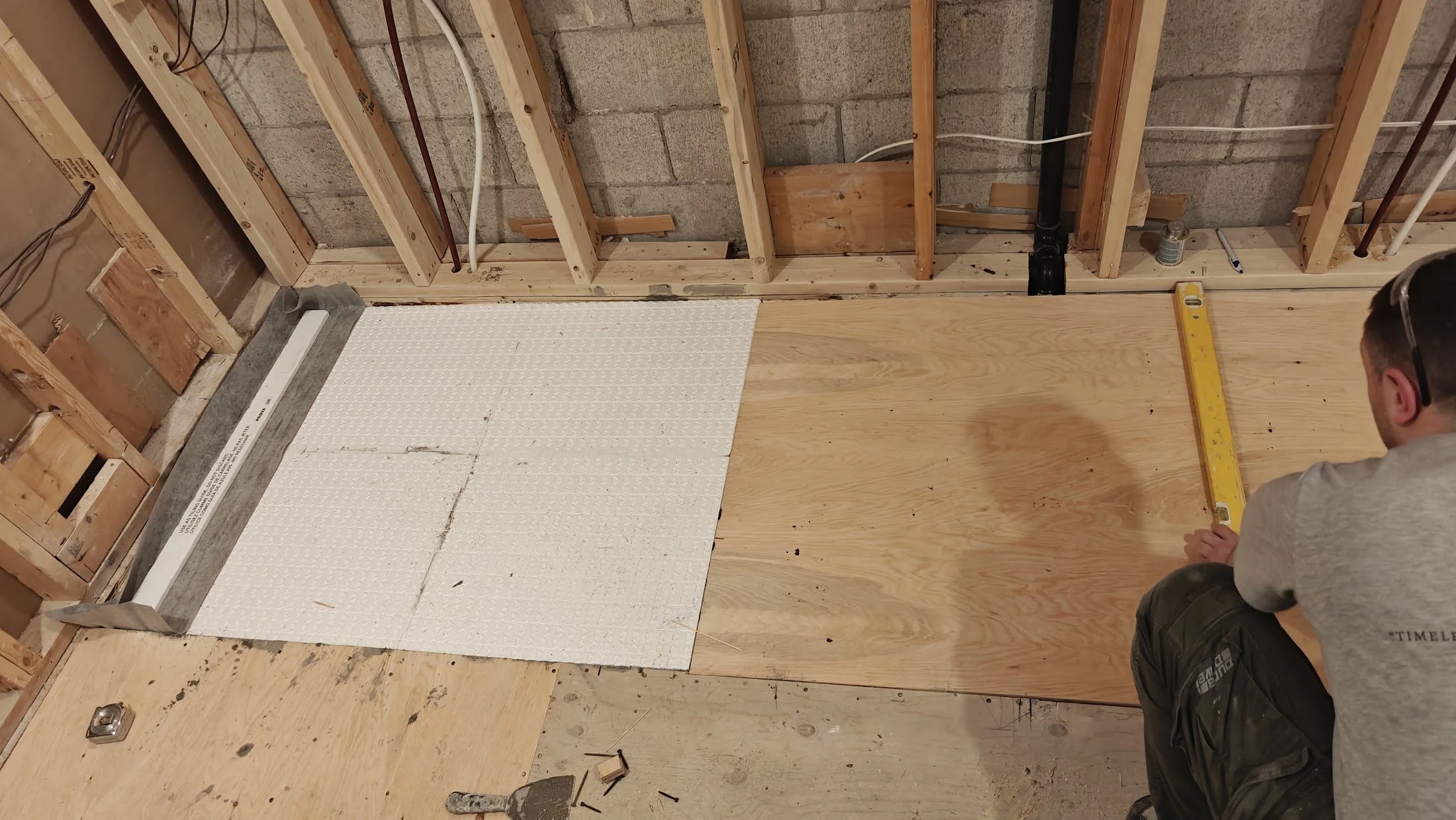 A person measuring a wooden floor with a yellow level in a partially constructed room. The room has exposed wall studs, some electrical wiring, and an unfinished concrete block wall. A section of the floor is being renovated, with some new wood floor