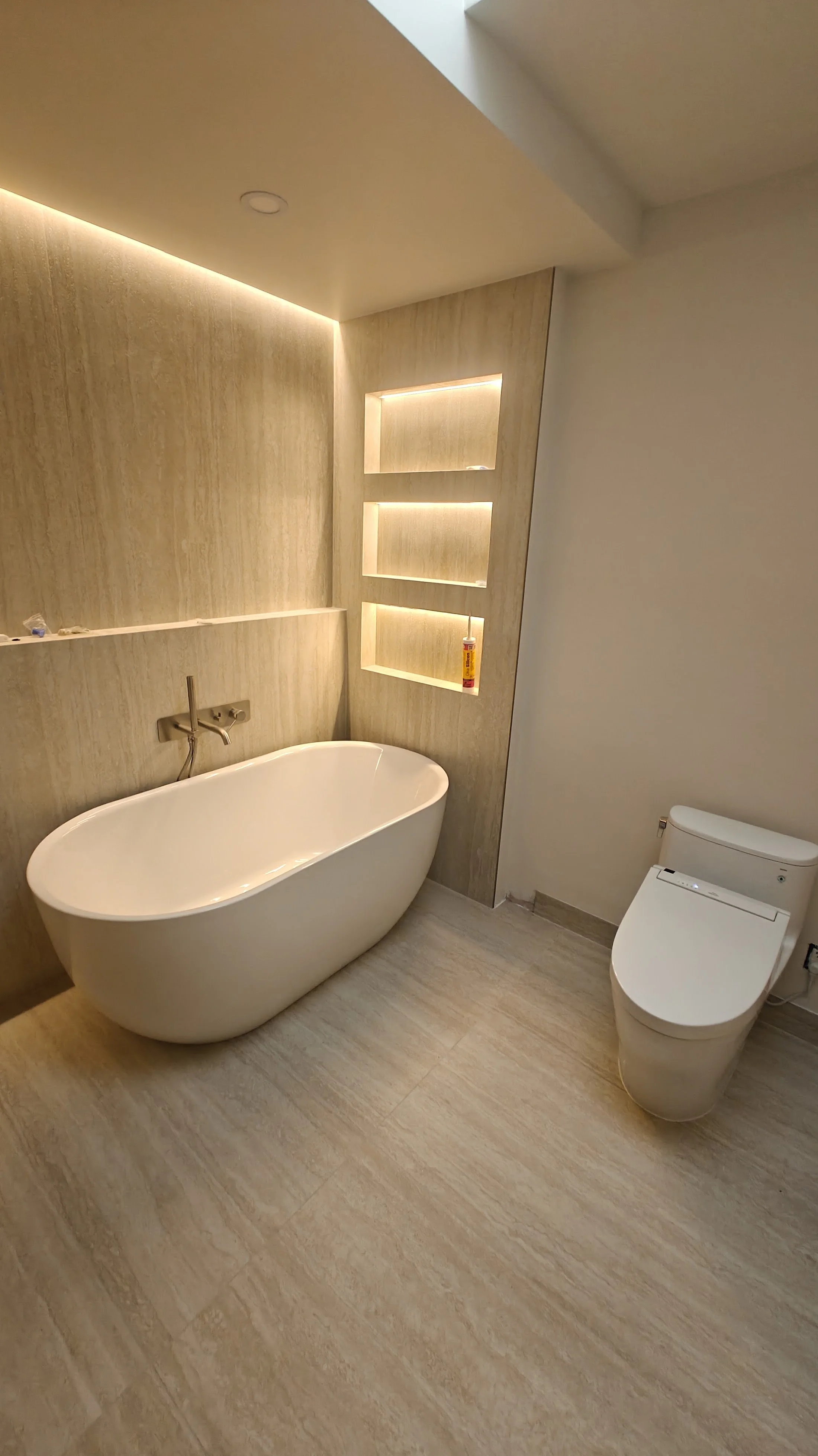 Modern bathroom with a white freestanding bathtub, a toilet, and built-in shelves with lighting on the wooden wall.