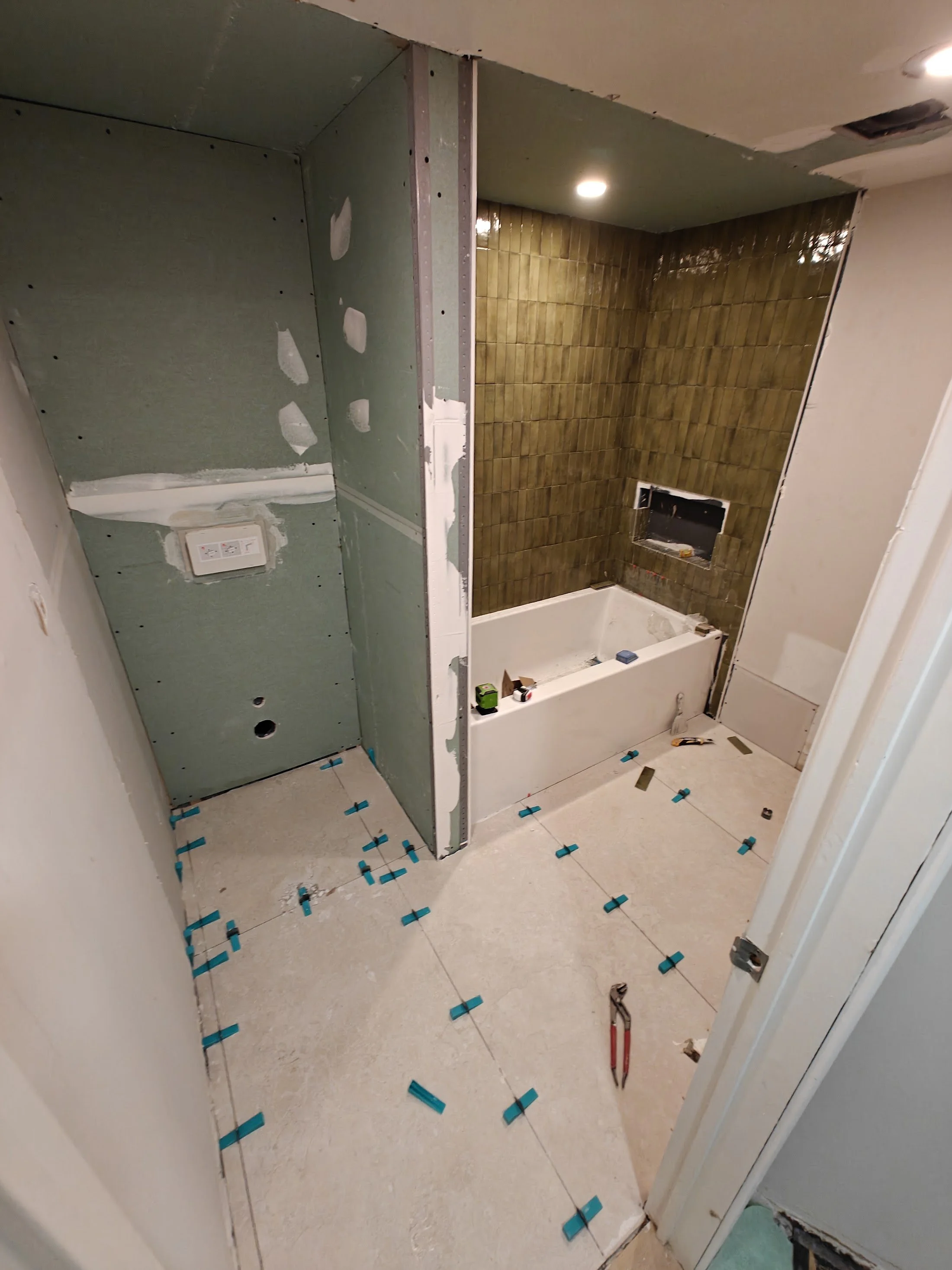 Bathroom under renovation with green drywall and brown tiled wall, floor tiles and construction tools visible.