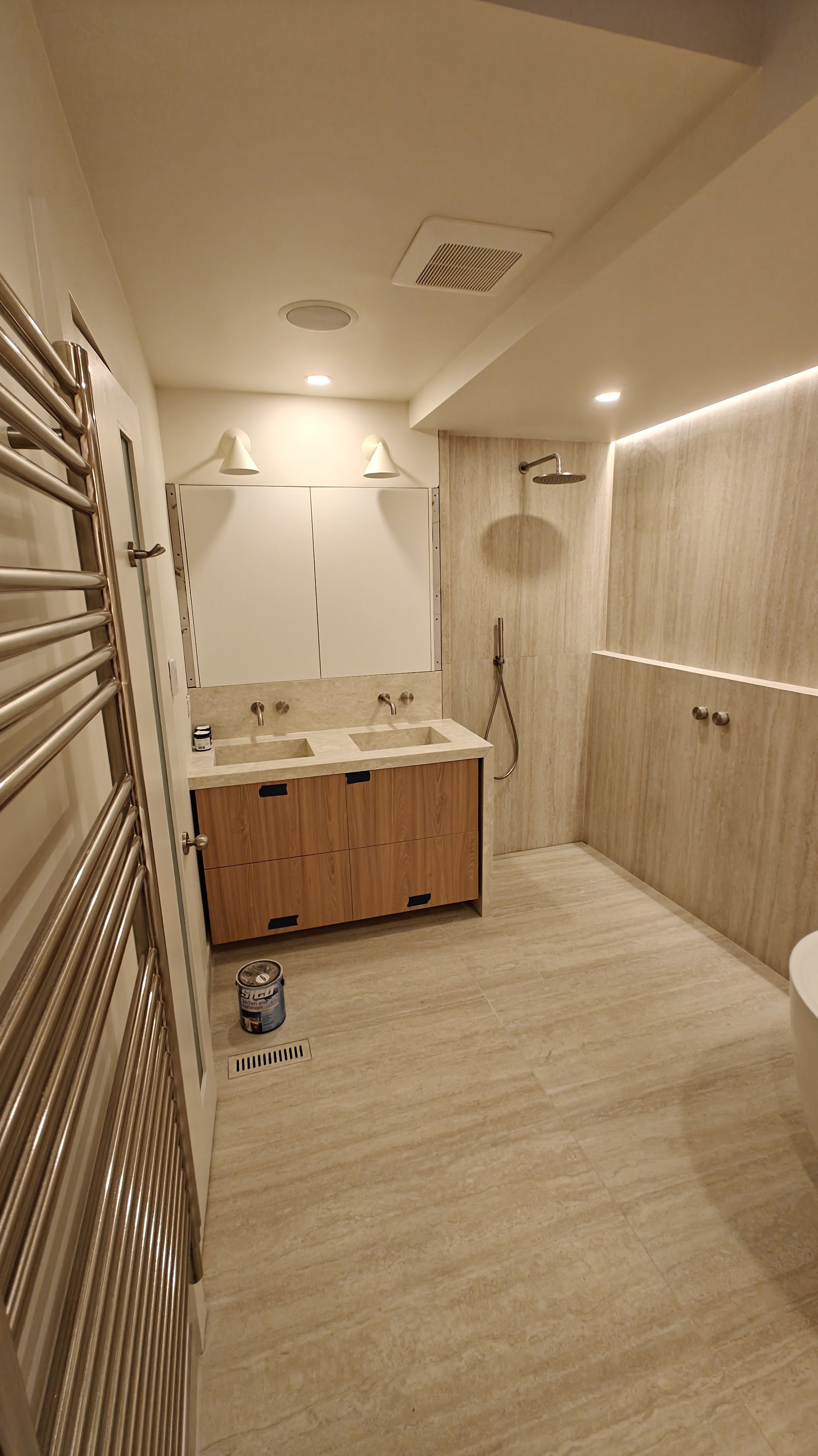 Modern bathroom with double sink vanity, wooden cabinet, wall-mounted showerhead in a beige marble shower area, heated towel rack, ceiling lights, and a window.
