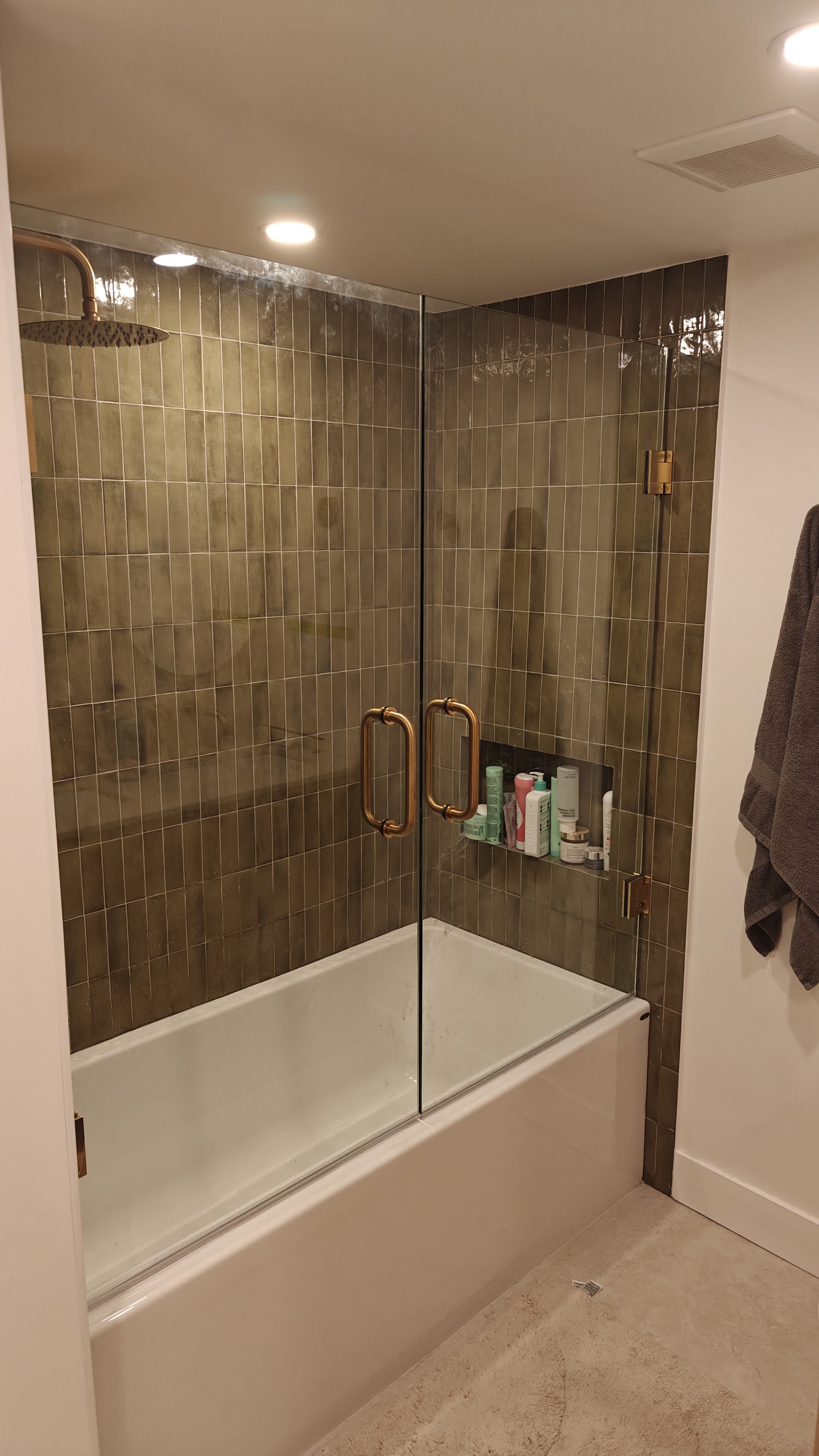 A glass-enclosed shower with brass handles, brown tile walls, a built-in shelf with toiletries, a ceiling light, and a towel hanging on a hook outside.