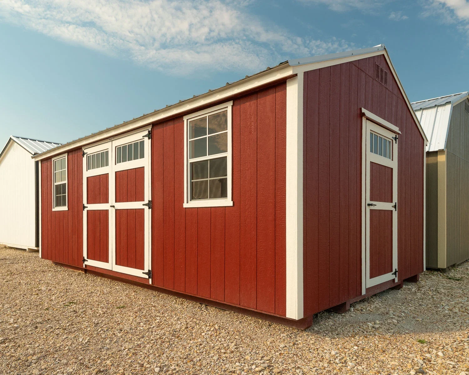 Red and white shed with windows and double doors, situated on a gravel surface under a partly cloudy sky.