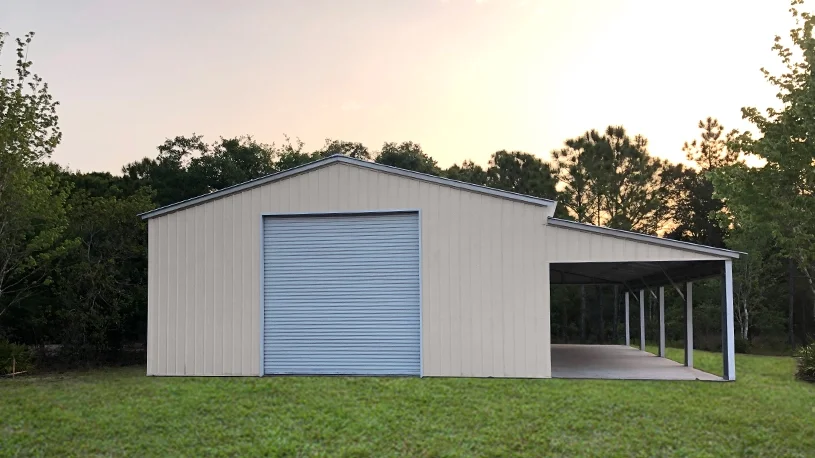 A white metal storage building with a roll-up garage door and a covered open area, on a grassy field with trees in the background during sunset.