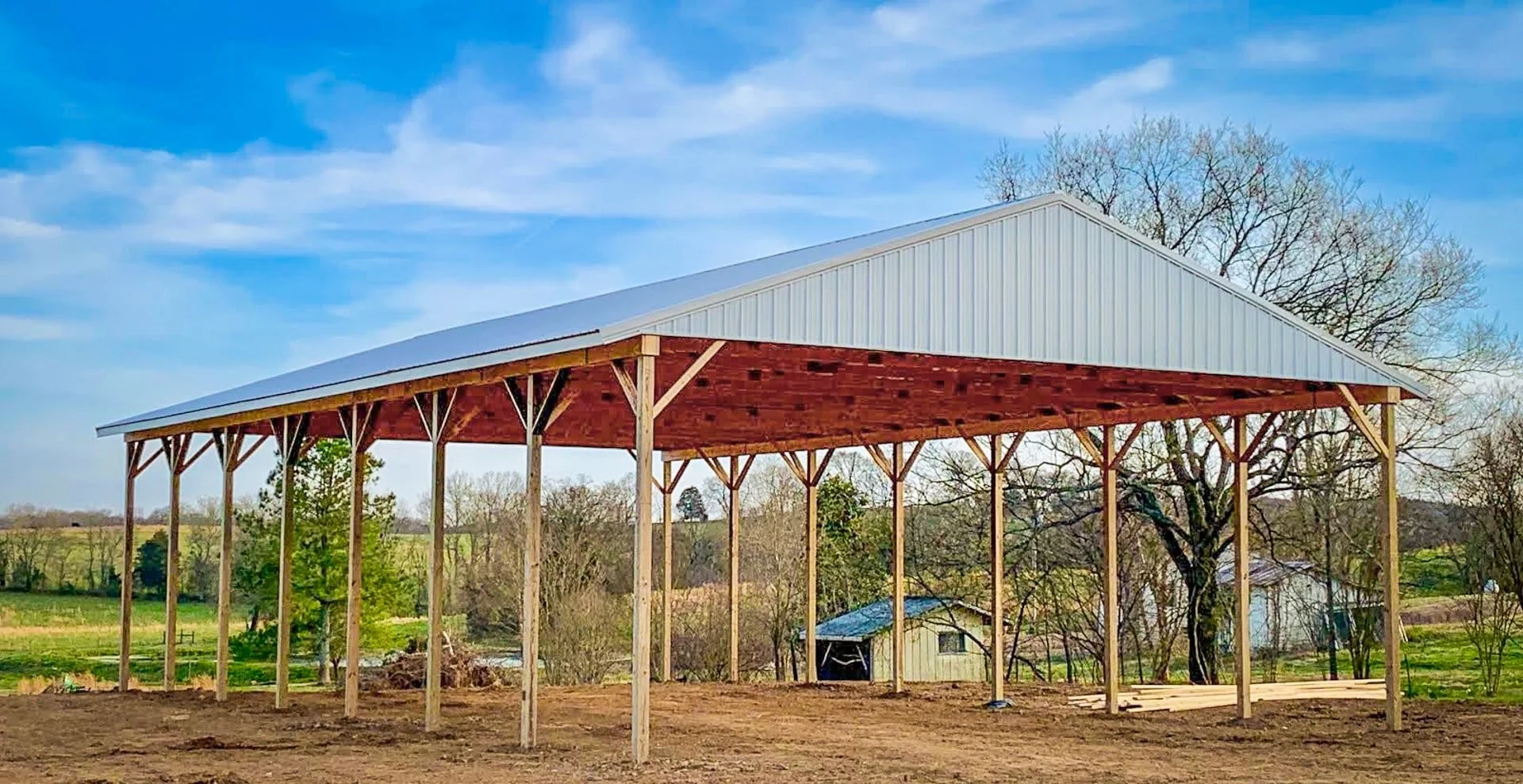 Under construction barn with metal roof and wooden supports during daytime, with trees and fields in the background.