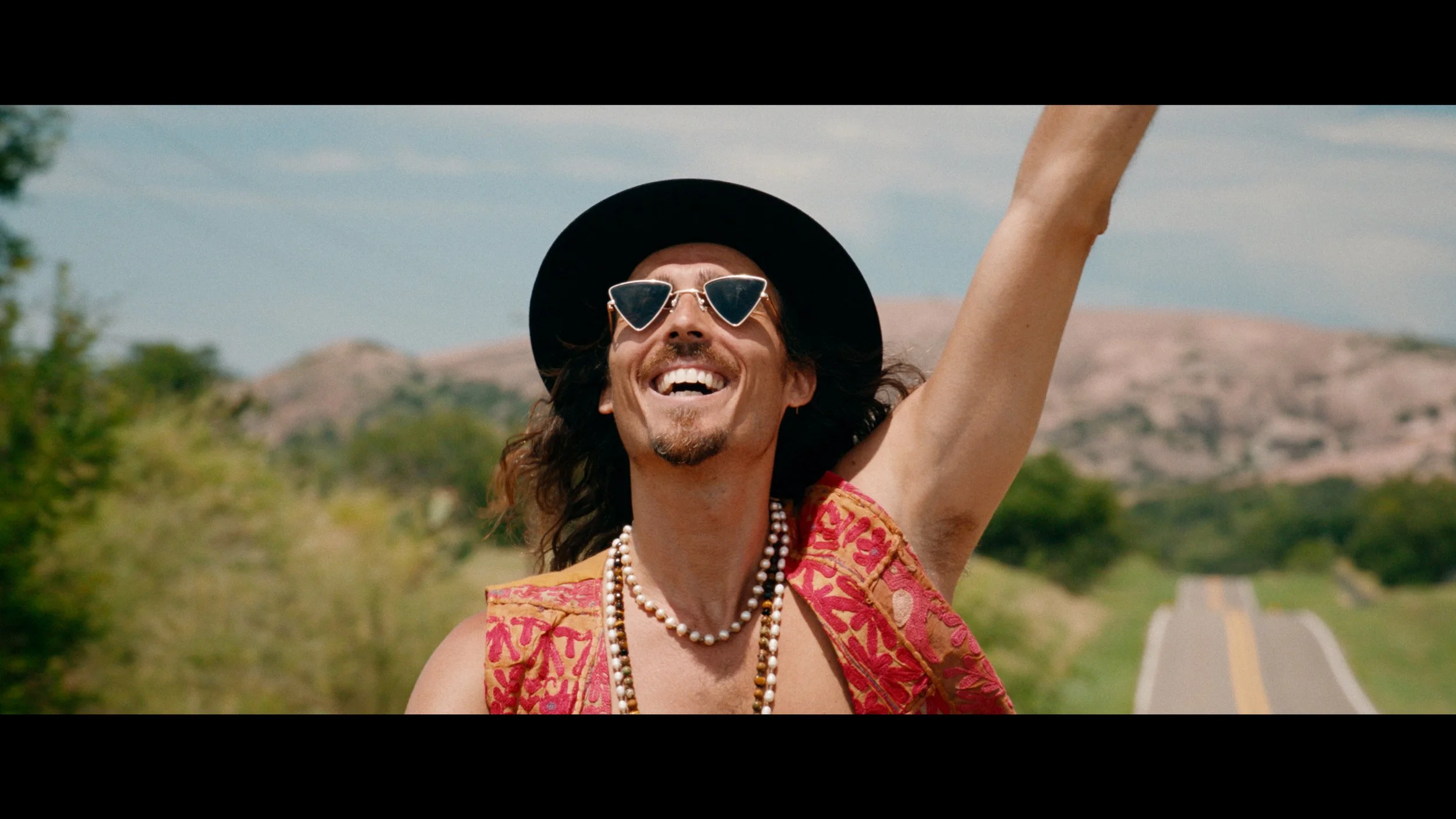 Dylan Townsend smiling and wearing sunglasses, a black wide-brimmed hat, a patterned red and pink top, and a pearl necklace, standing outdoors with hills and trees in the background.