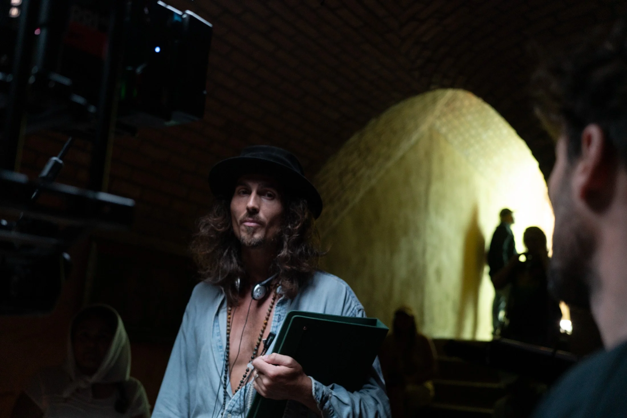 Dylan Townsend on set directing feature film wearing a hat and a blue shirt, is holding a tablet and a phone, engaging in conversation in a dimly lit space with an arched brick ceiling.