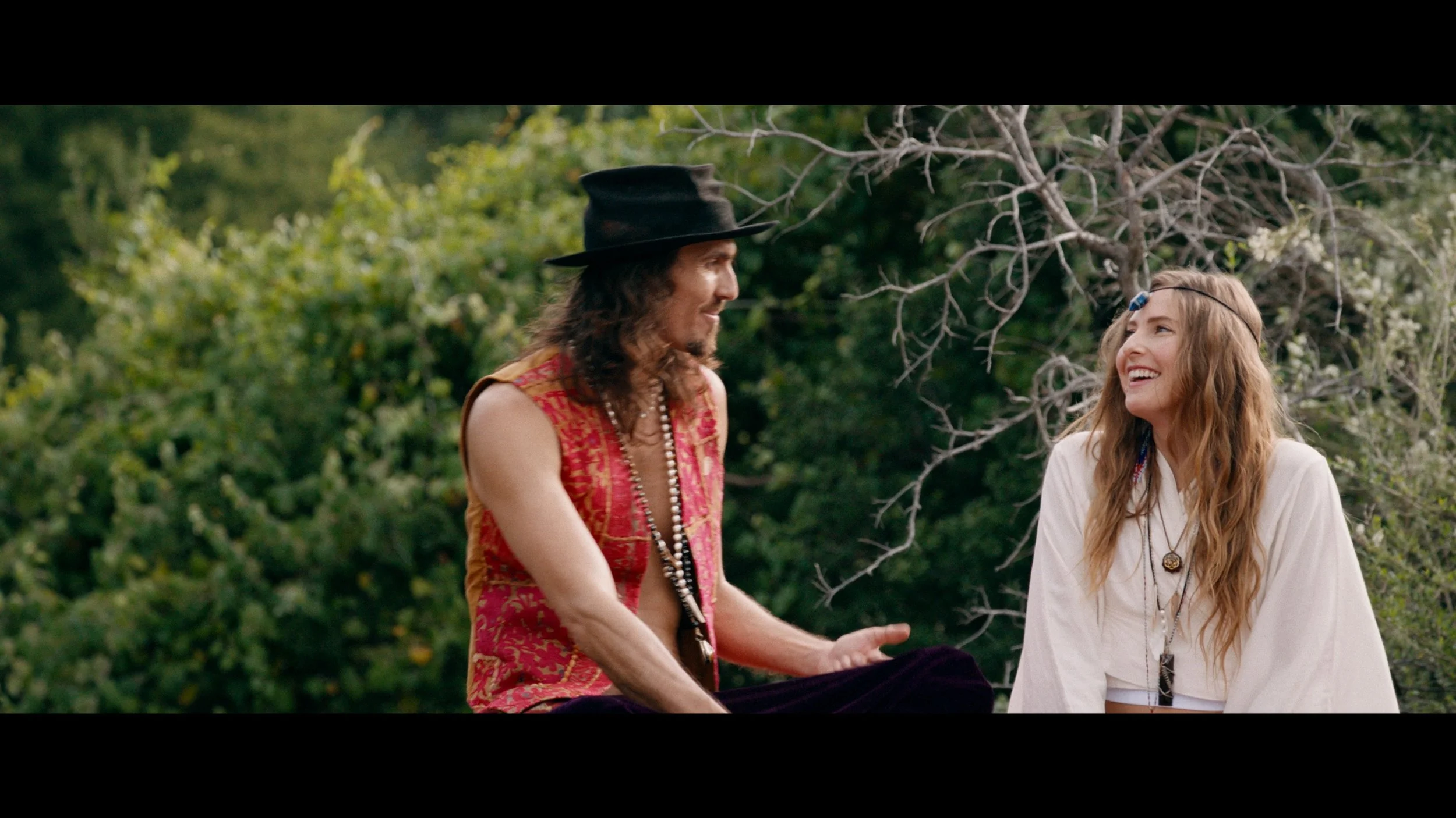 Dylan Townsend still from feature film with Two people, a man with long hair, a black hat, and a sleeveless vest, and a woman with long wavy hair and a headband, sitting and talking outdoors among greenery.