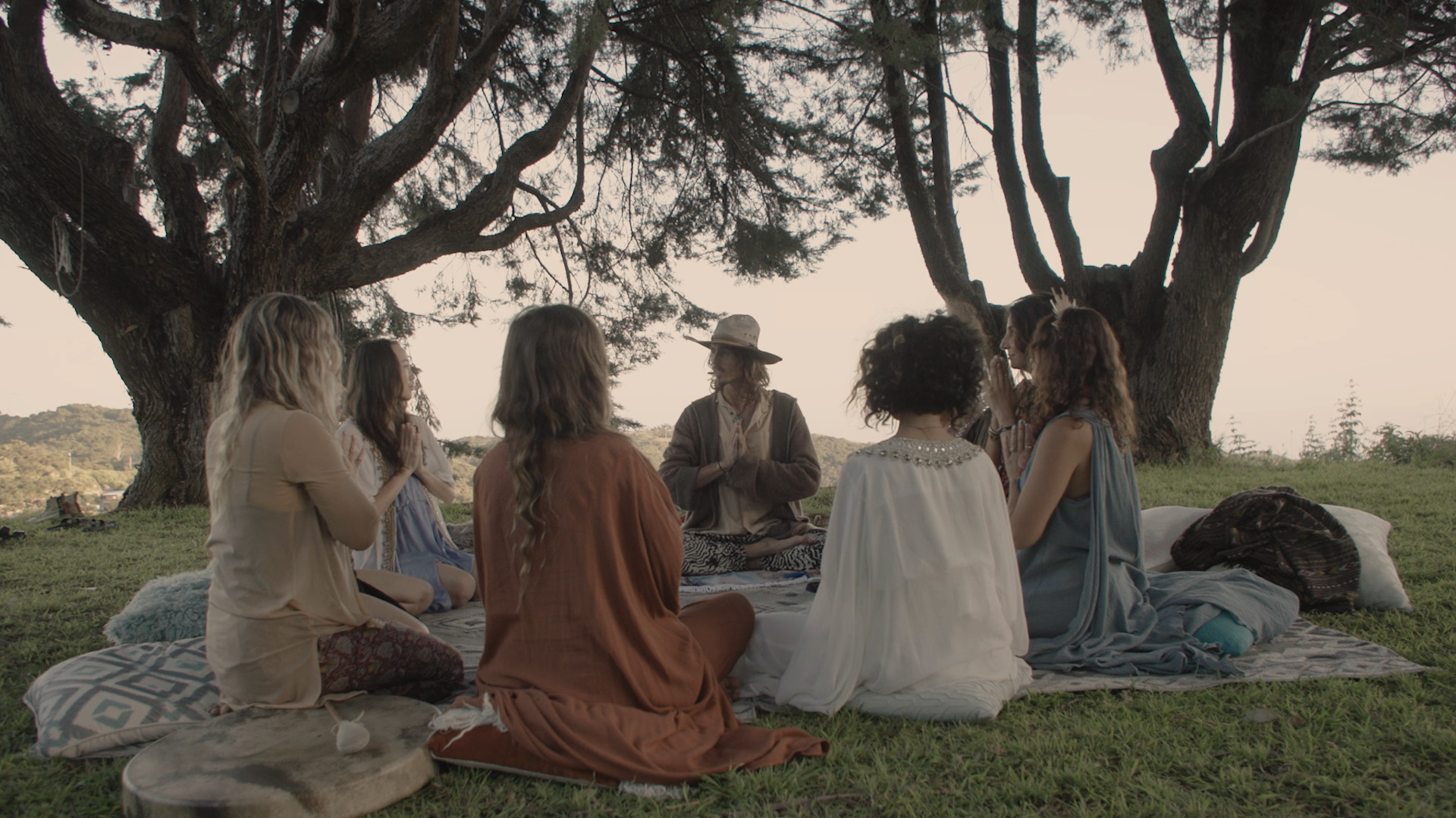 Dylan Townsend in 'Intentional' tv pilot. A group of women sitting cross-legged on blankets under a large tree, meditating outdoors during sunset.