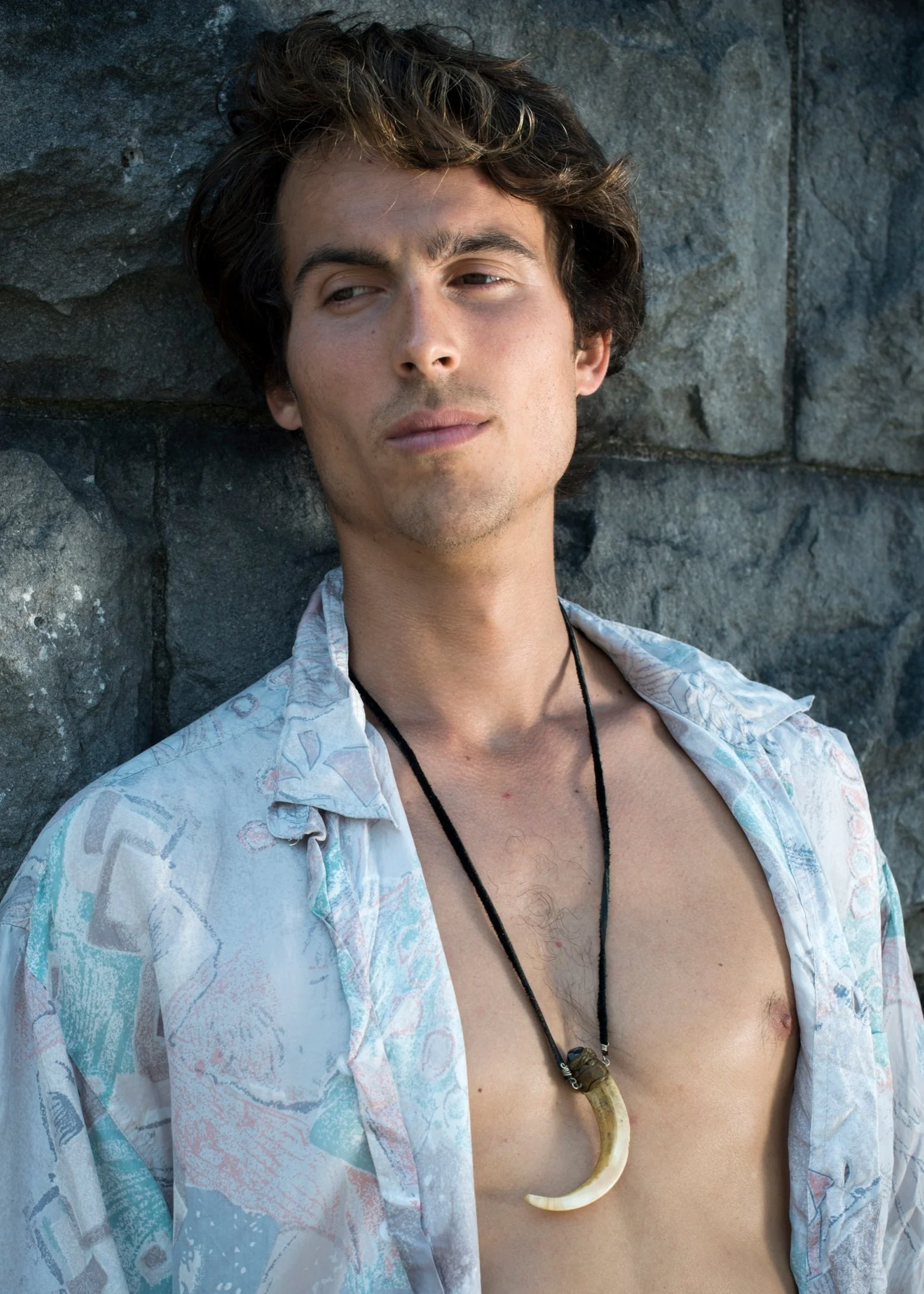 Young Dylan Townsend with tousled brown hair and light skin, wearing a partially open Hawaiian shirt and a black necklace with a horn pendant, leaning against a stone wall.