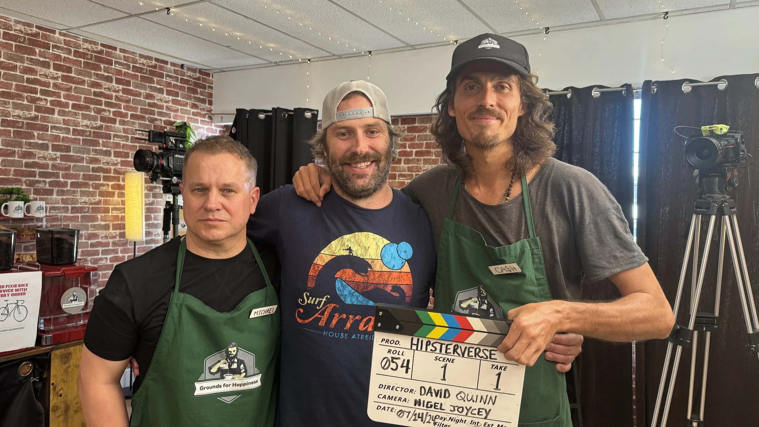 Dylan Townsend, David Laurence & M Coleman one holding a film clapperboard, in a room with brick and white walls, set up for filming, with cameras and books in the background. All are wearing casual clothes and green aprons, smiling at the camera.