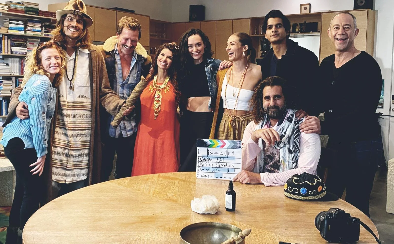 Dylan Townsend & Kirsten Sheridan on a film set, some dressed in bohemian and vintage style, gathered around a table with a clapperboard and filming equipment, behind a bookshelf in a room that seems to be a studio or office.