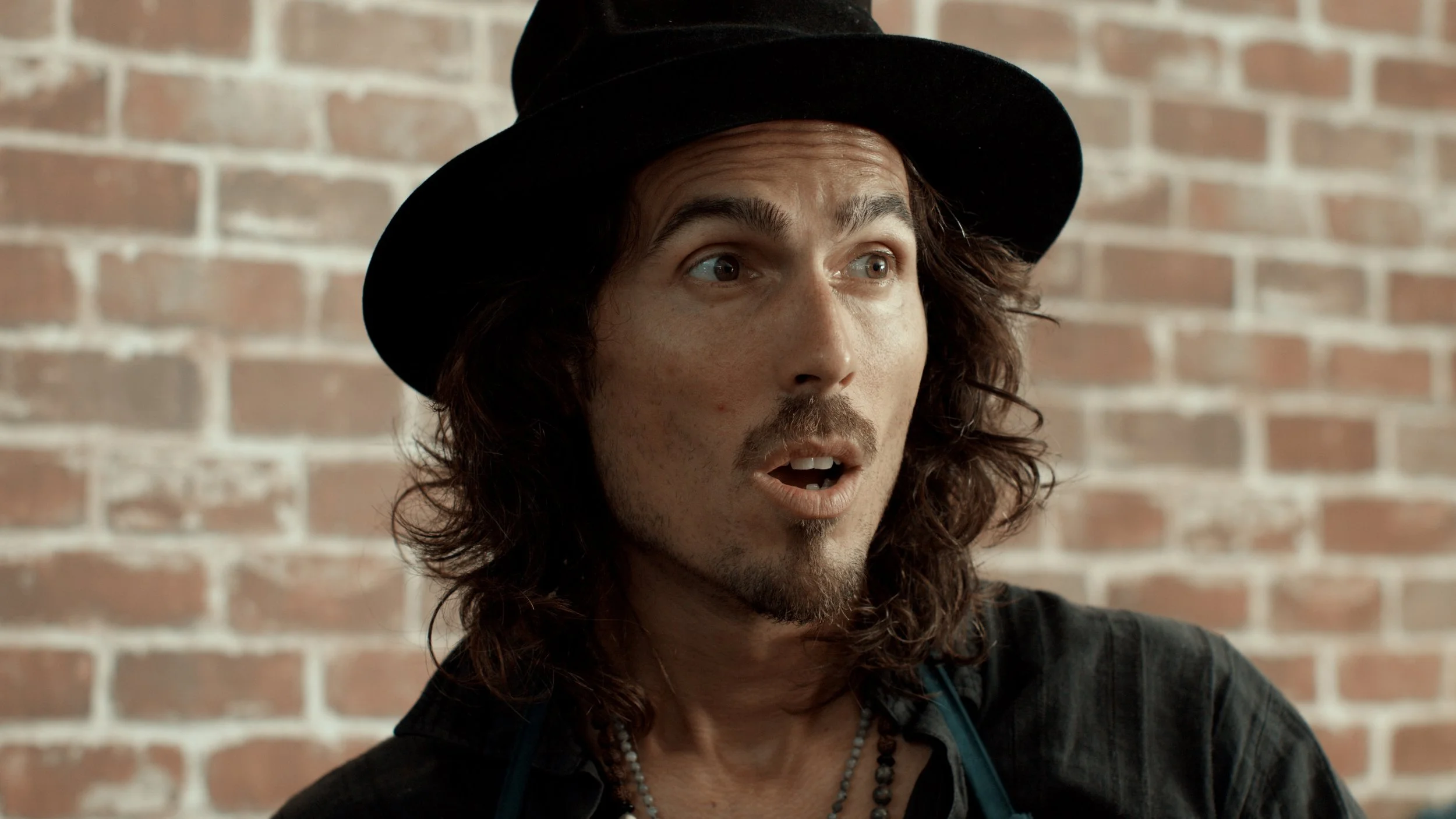 Dylan Townsend with long, curly brown hair, wearing a black hat, speaking or surprised, against a brick wall background.