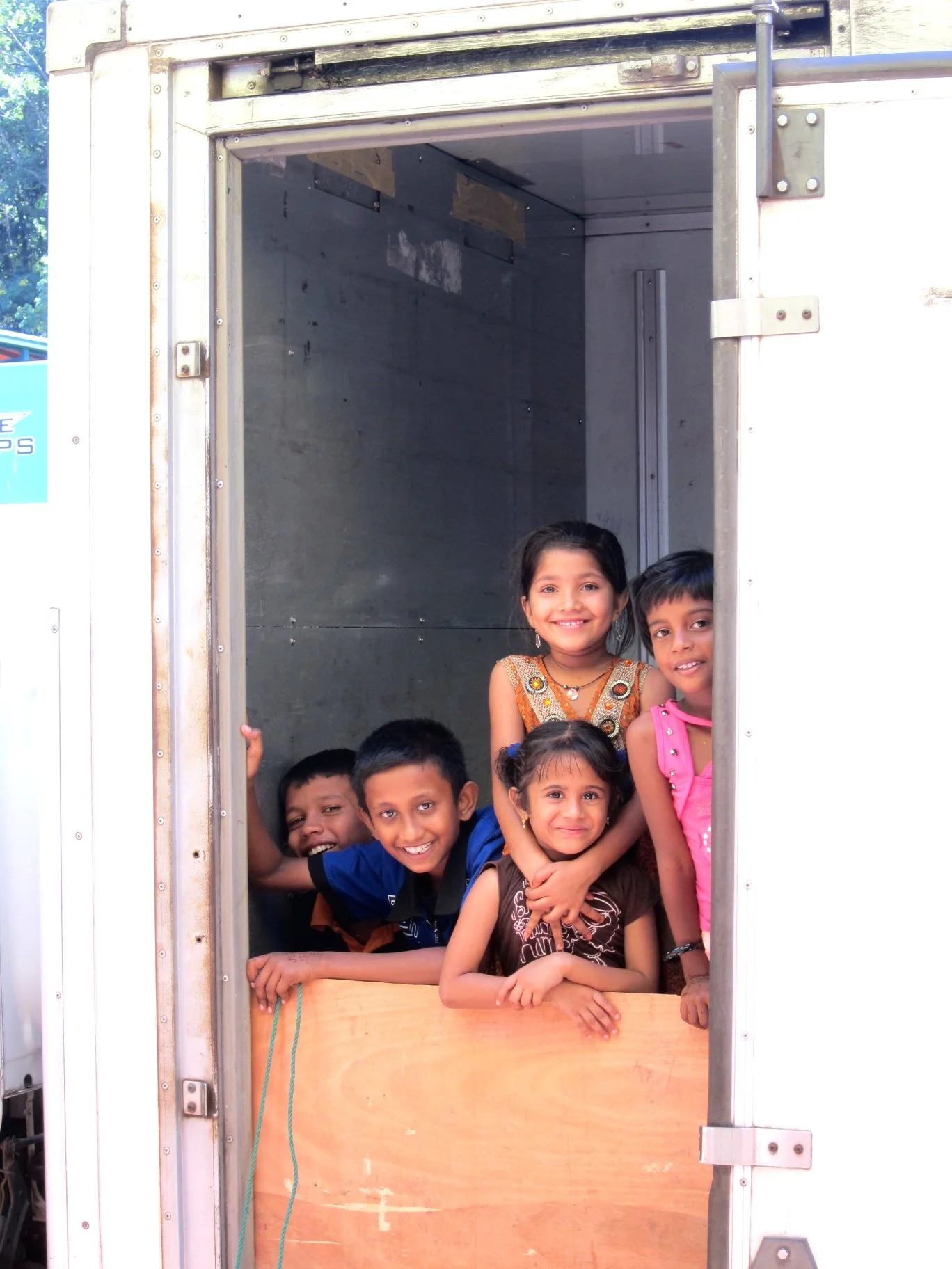 Children smiling and peeking out from the open door of a vehicle or box truck.