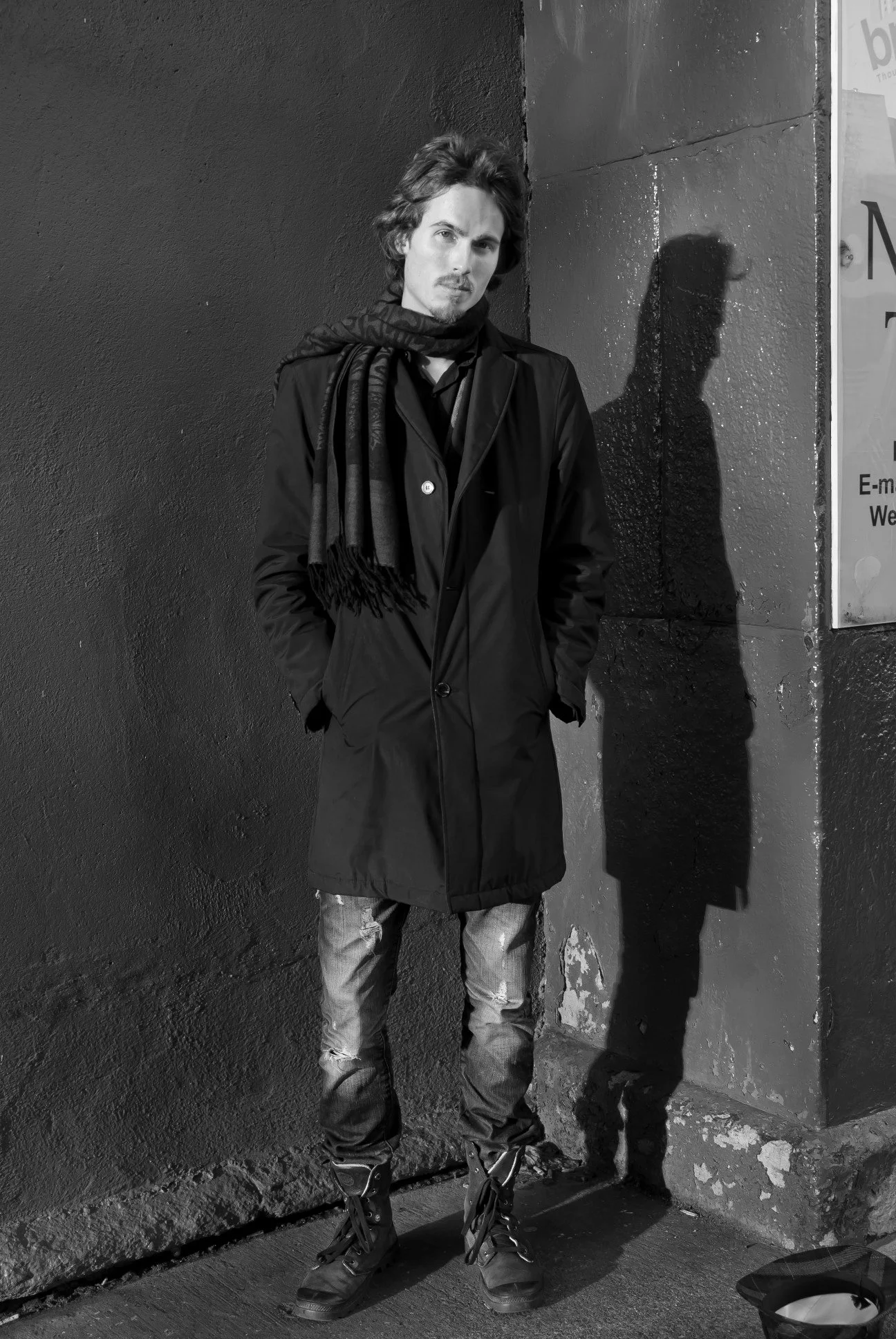 Dylan Townsend. With long hair and a mustache stands against a dark textured wall, wearing a long coat, jeans, and boots. His shadow and a part of a poster are visible on the wall. The photo is black and white.